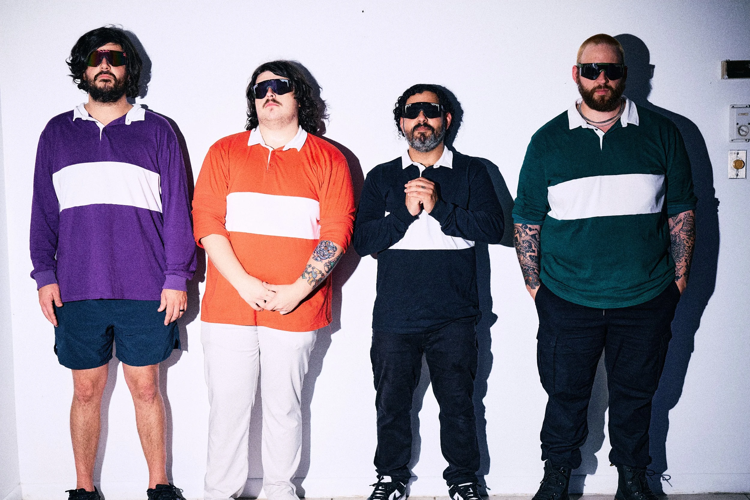 Four men standing against a plain white background, all wearing sunglasses and colorful shirts with white horizontal stripes, with tattoos visible on some of their arms.