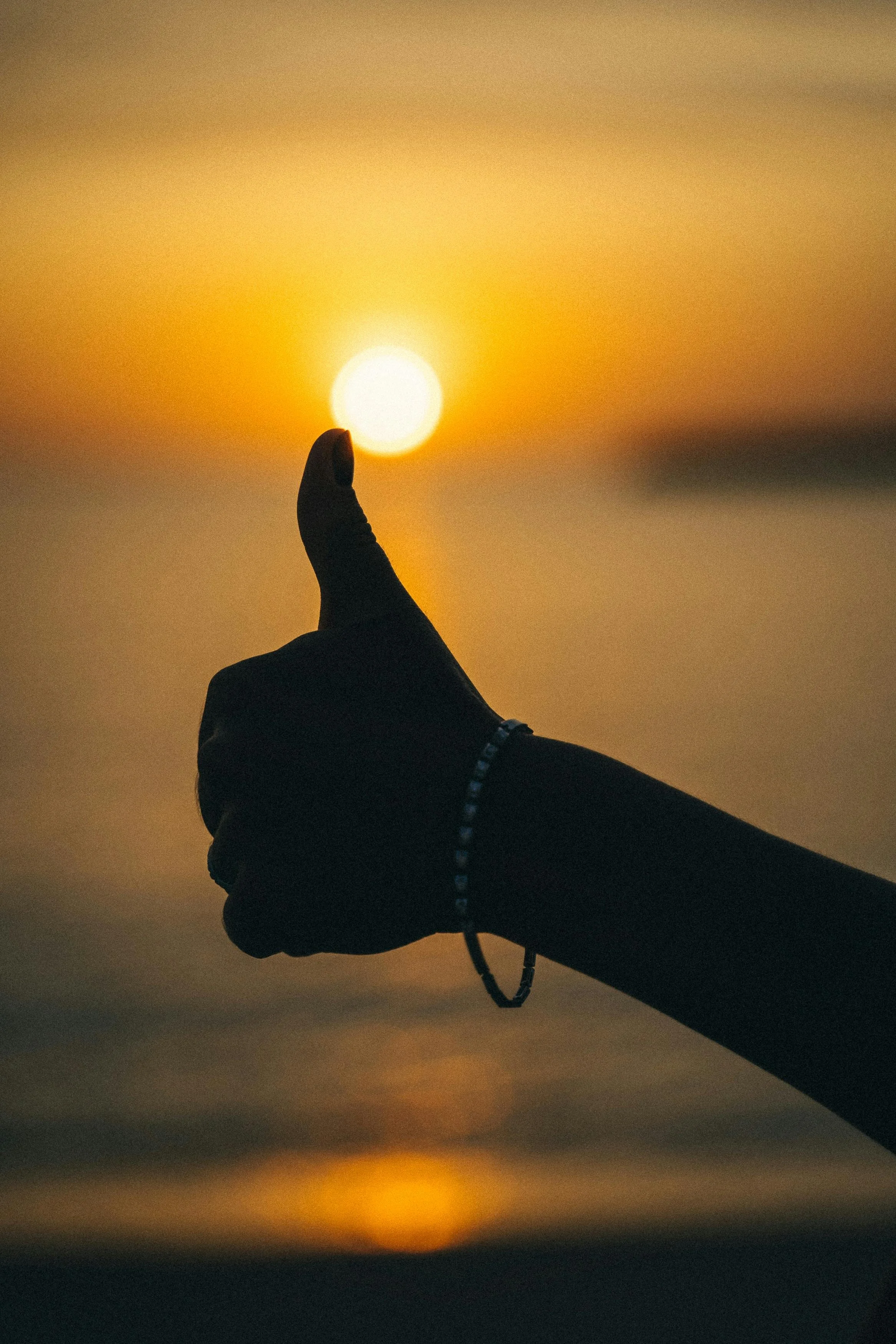 A person’s hand with a bracelet making a thumbs-up gesture, held up against a sunset background over water.