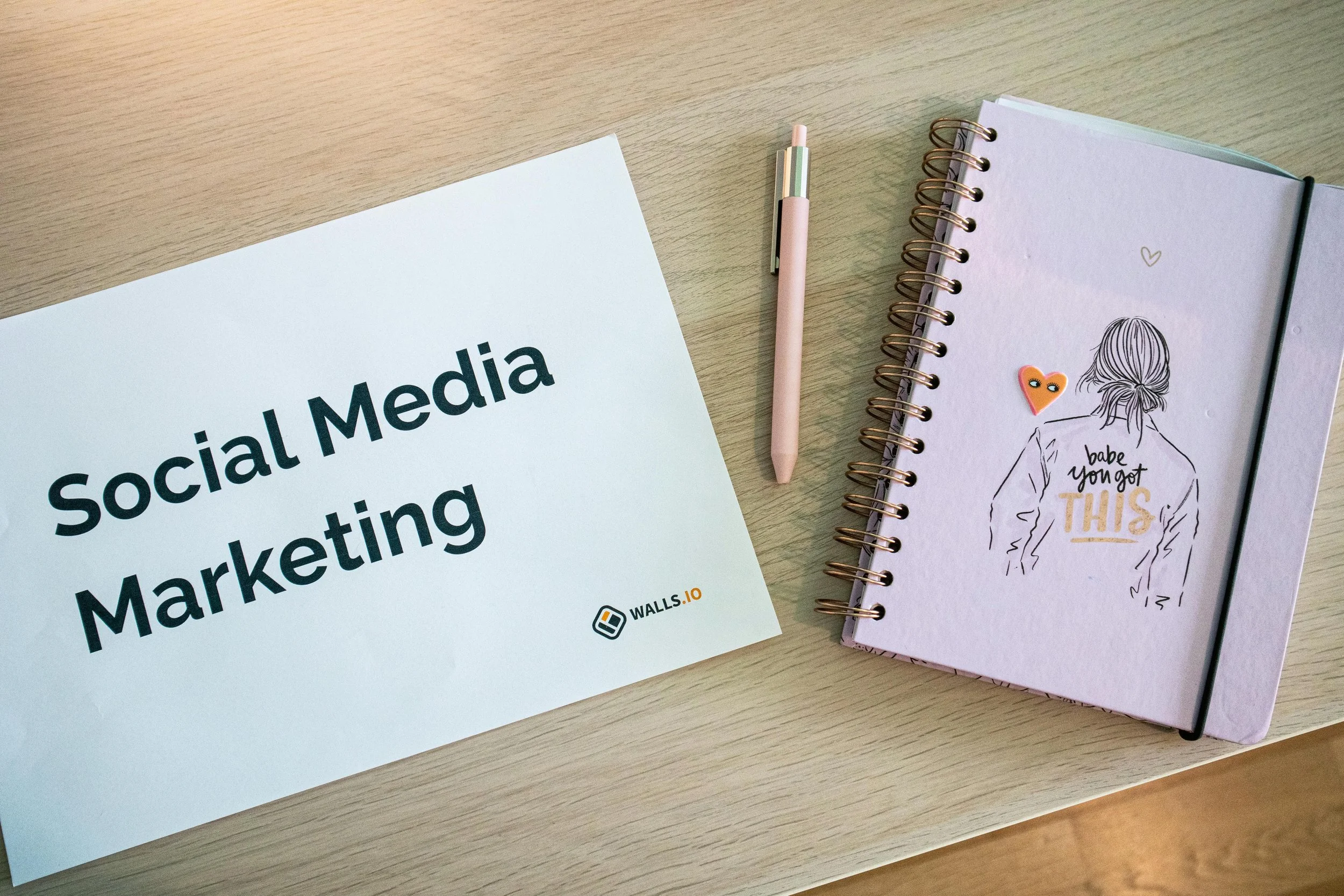 A printed sheet titled 'Social Media Marketing' next to a pink pen, and an open notebook with a line drawing of a woman on the cover with text 'babe you got THIS' and heart stickers on a wooden table.