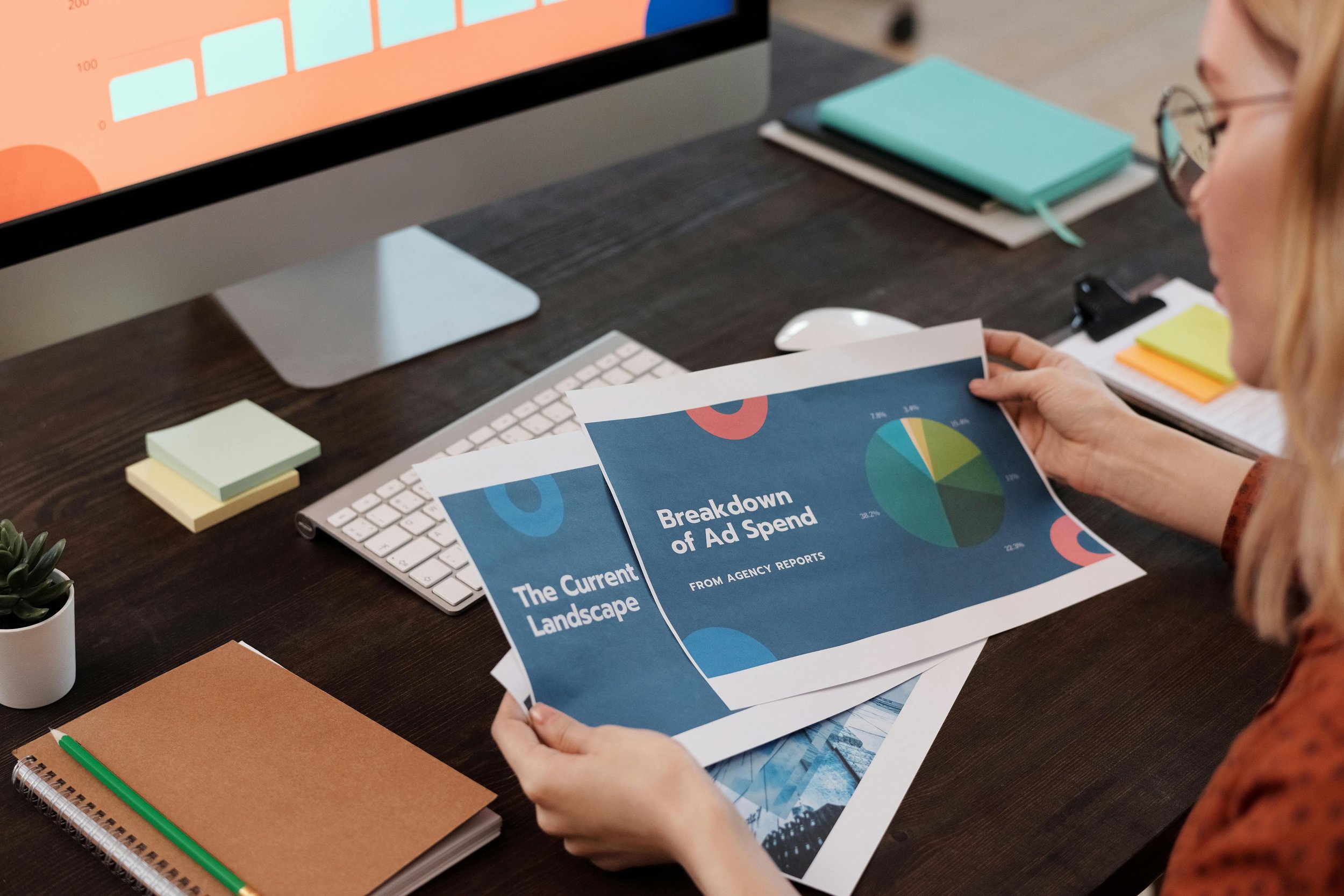 Woman reviewing printed reports on ad spend at her desk with a desktop computer, notebooks, sticky notes, and a plant.