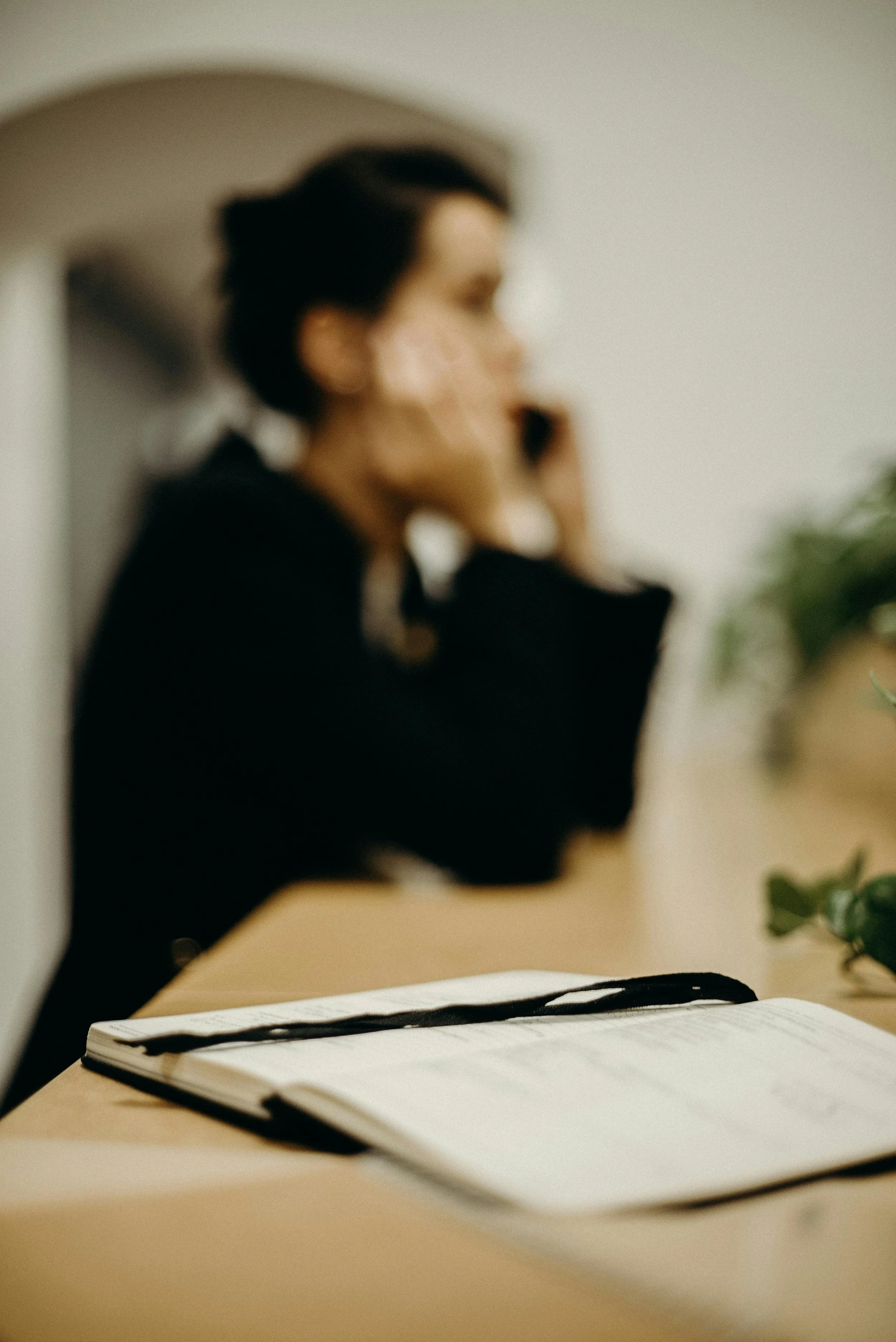 Open notebook with black ribbon on desk, woman in black shirt in background, blurred, talking on phone.
