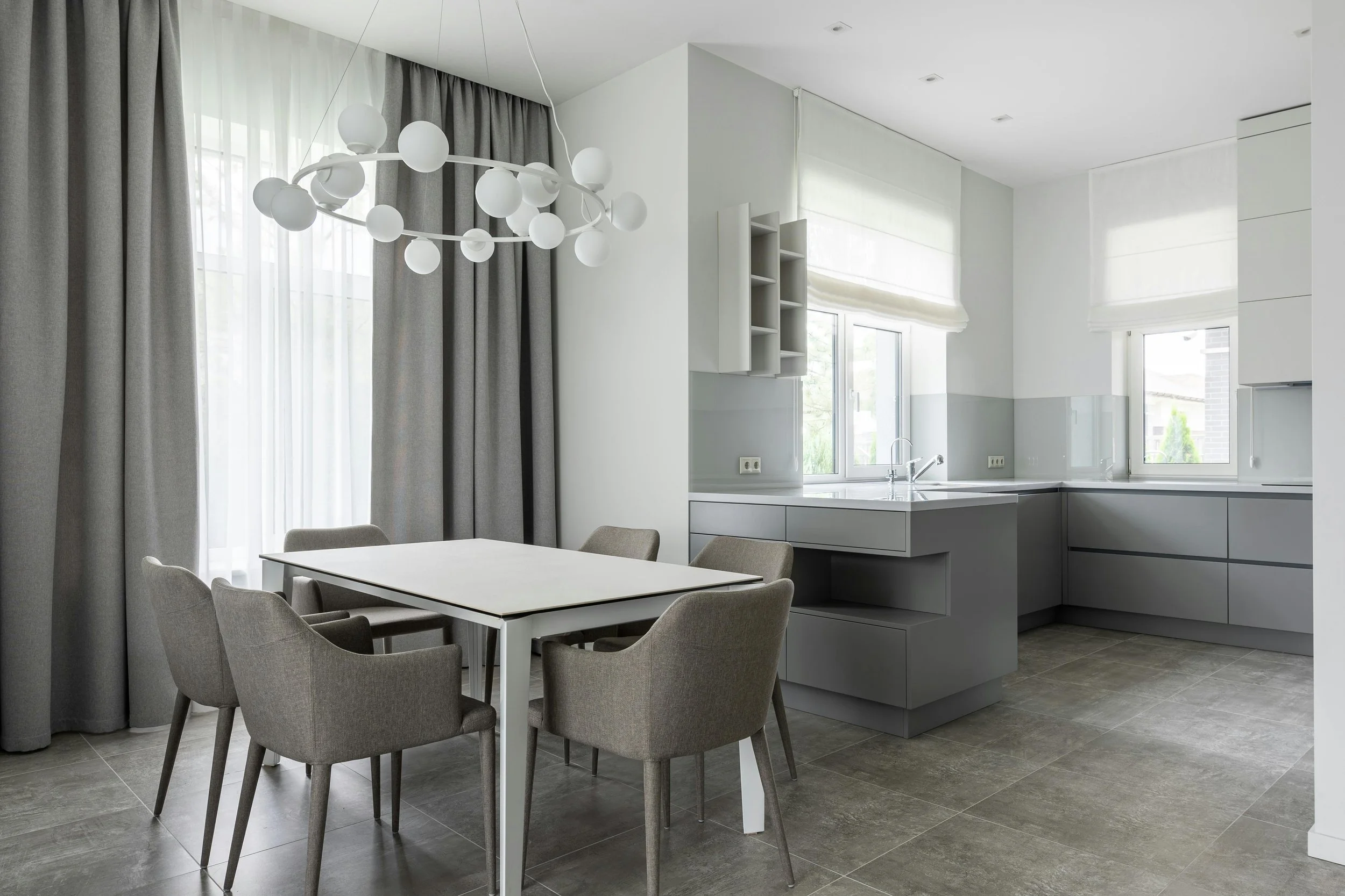 Modern kitchen and dining area with gray cabinets, white countertops, and a white dining table with six gray chairs. Large windows with white Roman shades and gray curtains, and a contemporary chandelier with white globe lights.