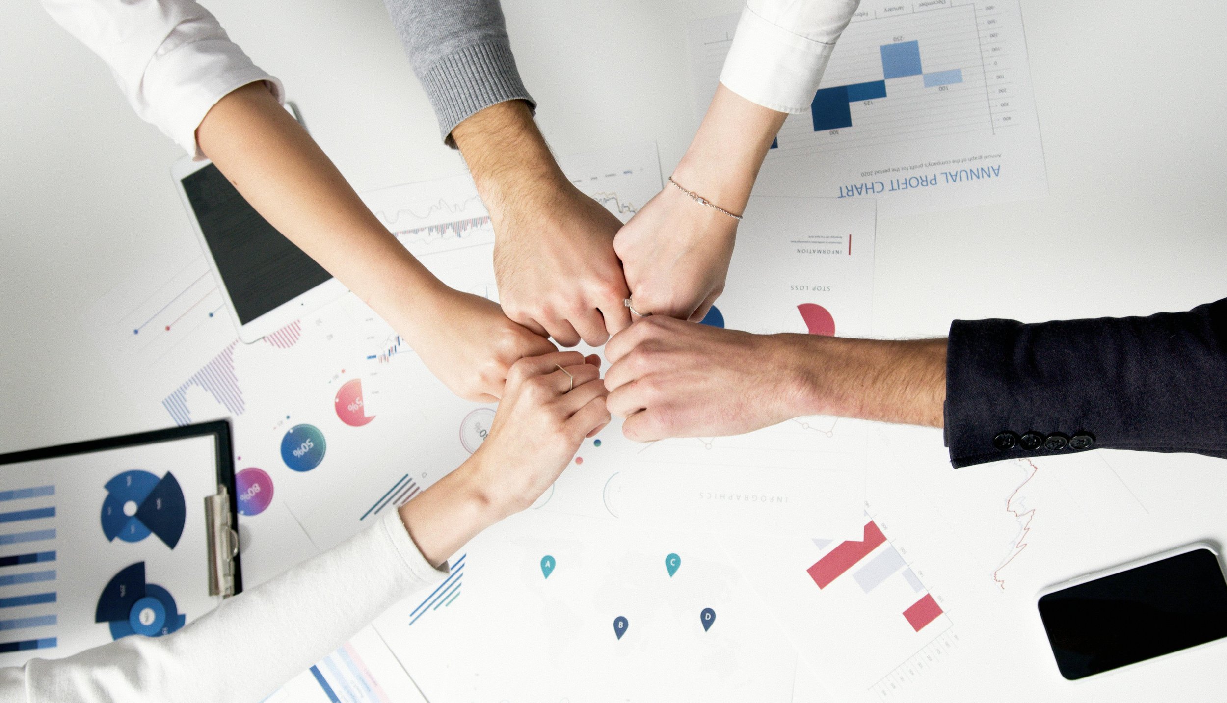 Four people placing their fists together in a team gesture over a desk with charts and graphs, including bar graphs and pie charts, and a smartphone.