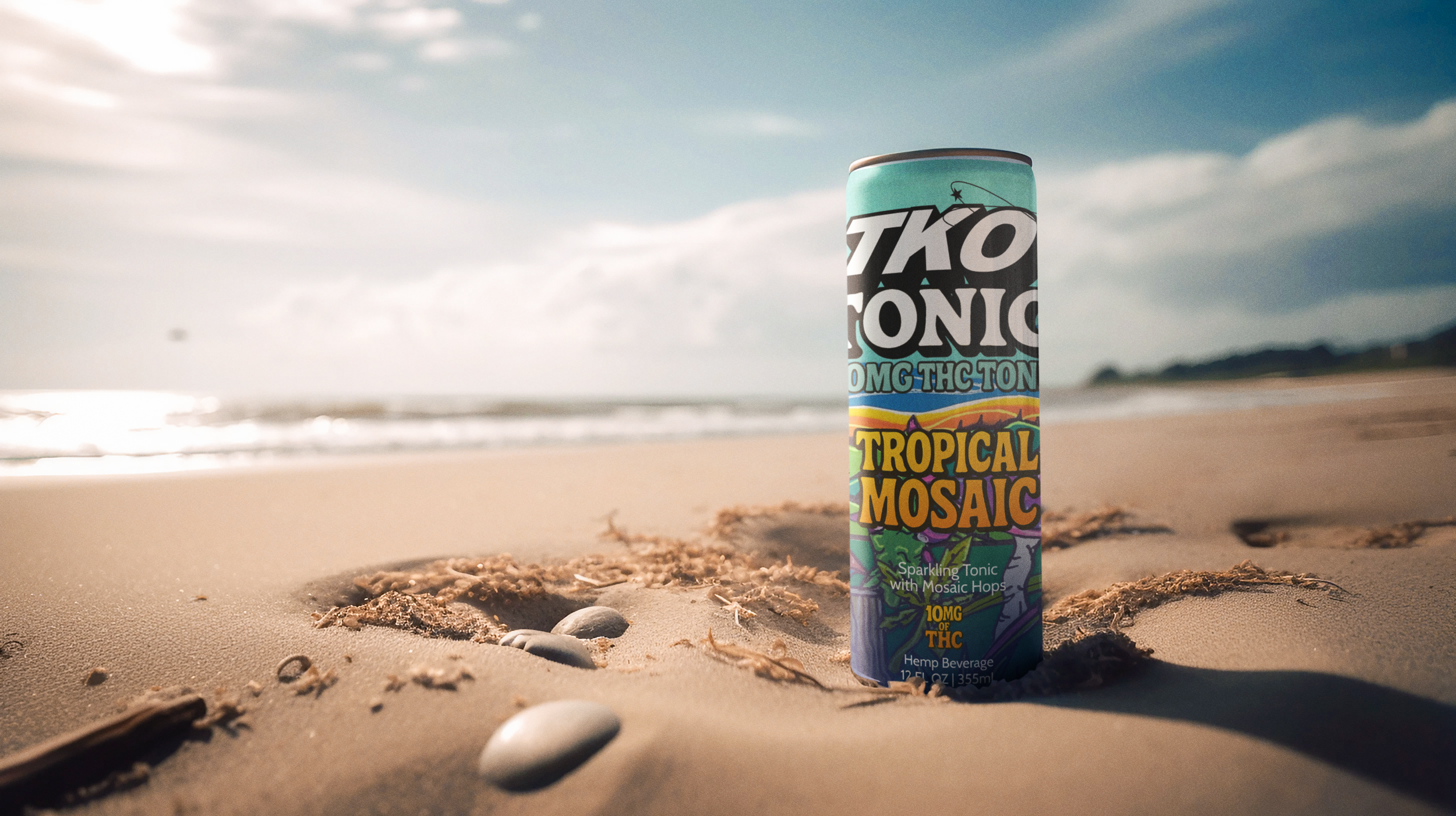 A can of Tropical Mosaic sparkling tonic with mosaic hops standing upright on sandy beach, with seashells and seaweed around, ocean waves in the background, under a partly cloudy sky.