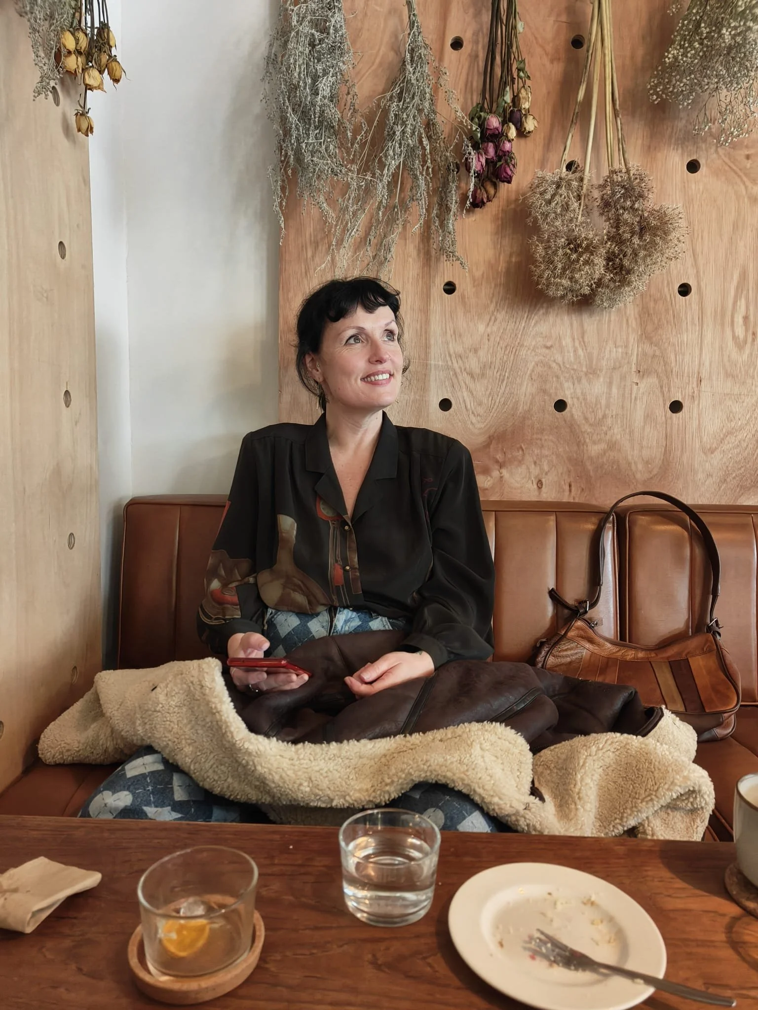 A woman sitting on a brown leather bench in a cozy cafe, holding a red phone, with dried flowers hanging on a wooden wall behind her, a leather jacket and bag beside her, a glass of water, a plate with some crumbs and a fork in front of her.