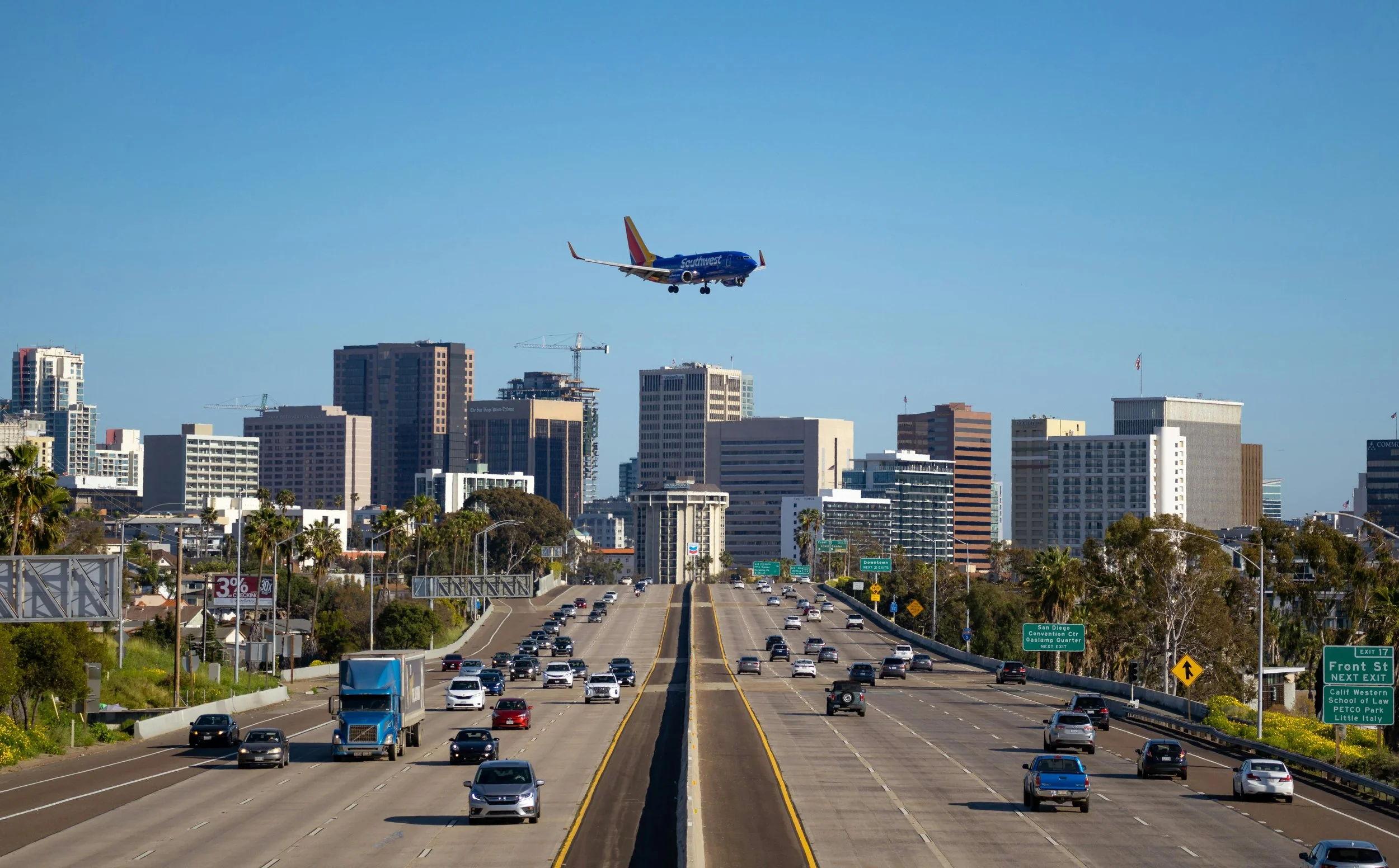 A busy highway leading to a city skyline with high-rise buildings, and an airplane flying overhead in clear blue skies.