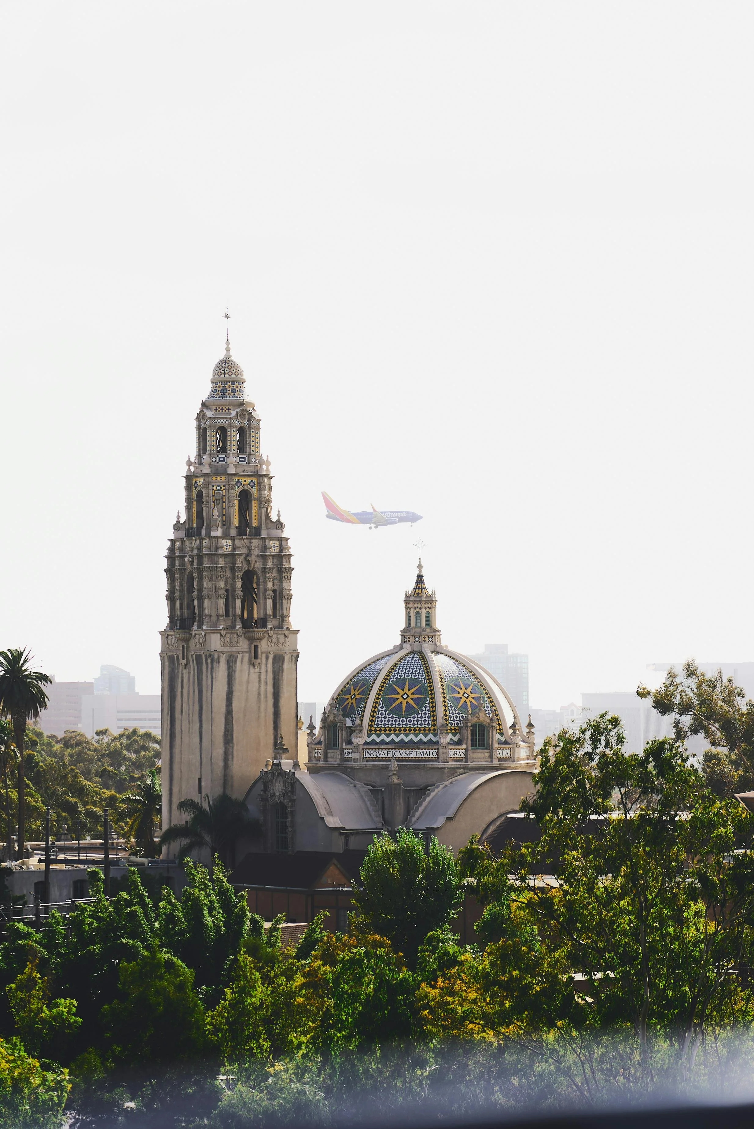 A cityscape featuring a historic church with a tall bell tower and colorful tiled dome, surrounded by lush trees, with an airplane flying in the sky in the background.