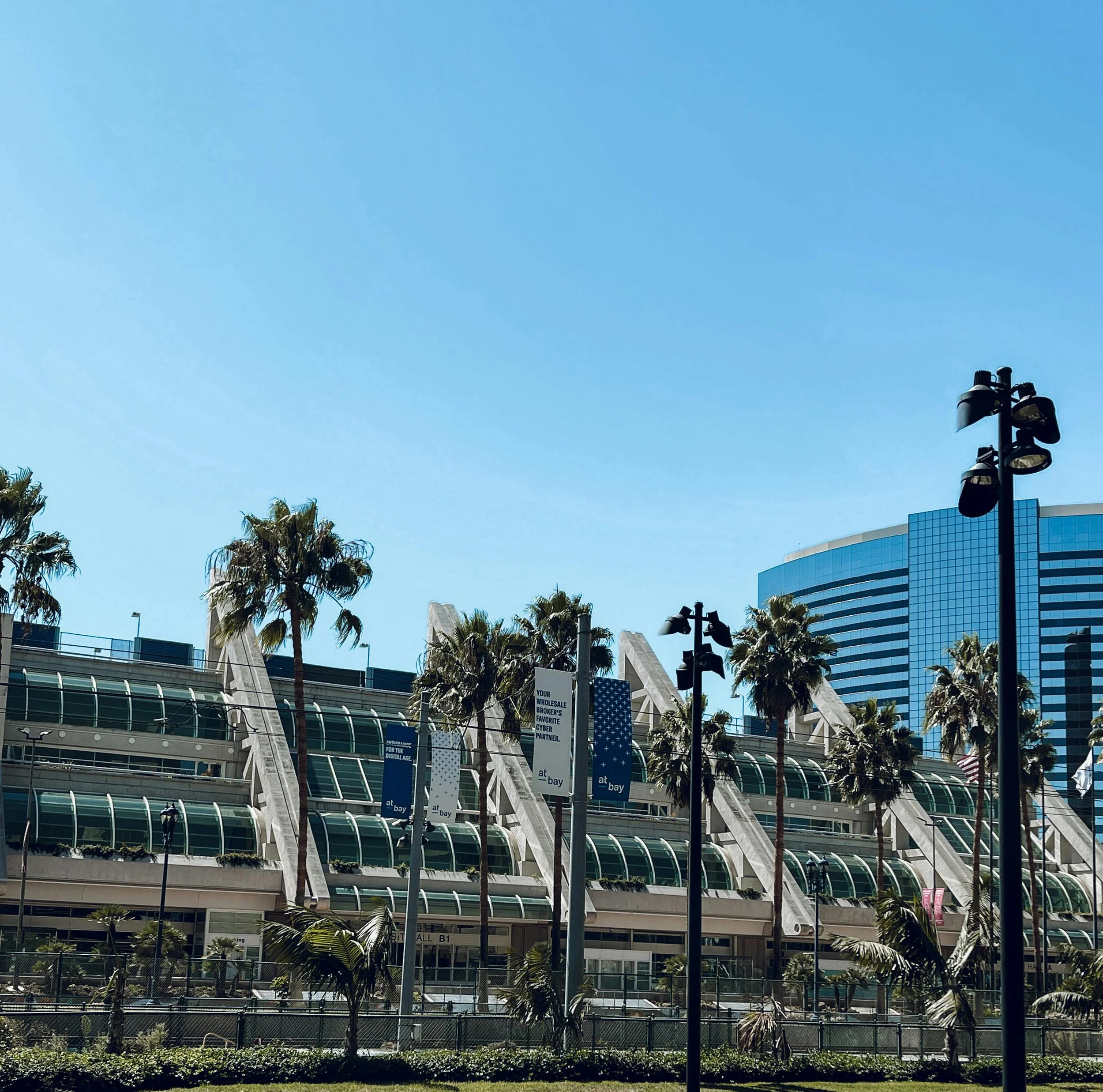 Modern convention center with glass-roofed walkways, surrounded by palm trees, with high-rise buildings in the background, under a clear blue sky.
