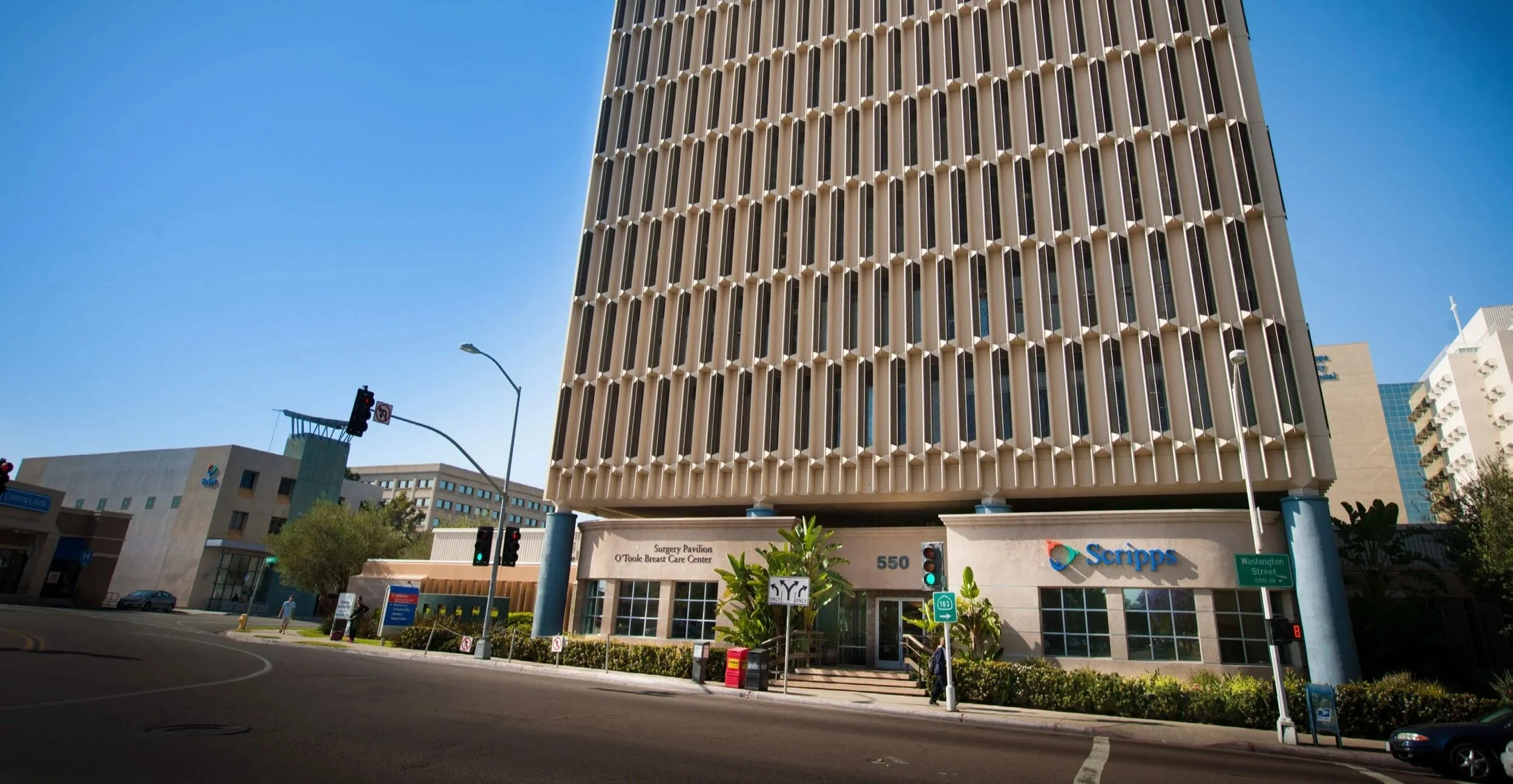 A tall office building with a beige, grid-like facade, located on a city street corner. The building has signage for Scripps and a surgery pavilion below. There are traffic lights, street signs, and a few pedestrians nearby.