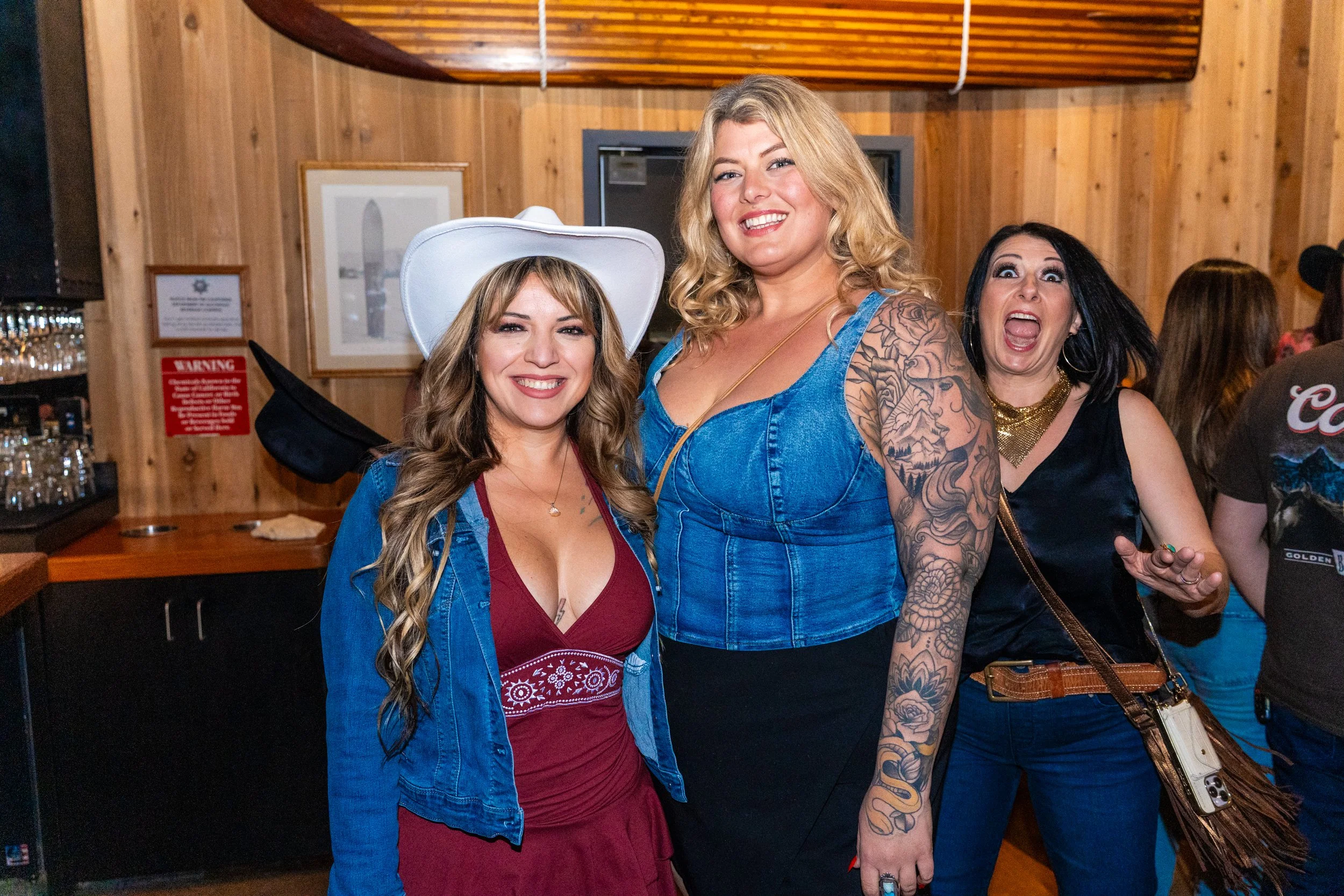 Three women at a social gathering in a wooden-paneled room. The woman on the left wears a white cowboy hat and a denim jacket over a red dress, smiling at the camera. The woman in the middle wears a blue sleeveless top, and has blonde hair and tattoo