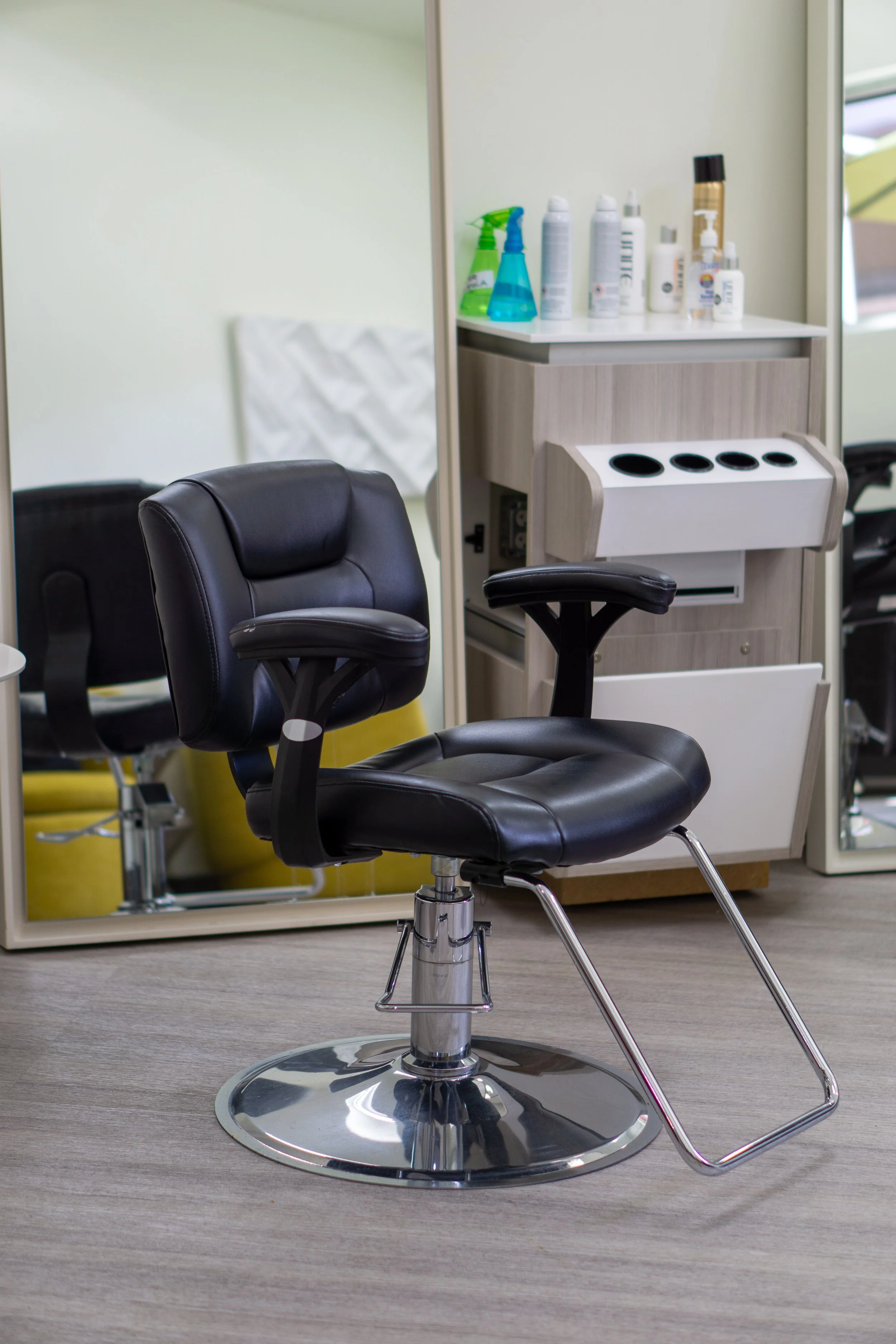 A black salon chair with a footrest, in front of a mirror and a cabinet with grooming products and sprays.