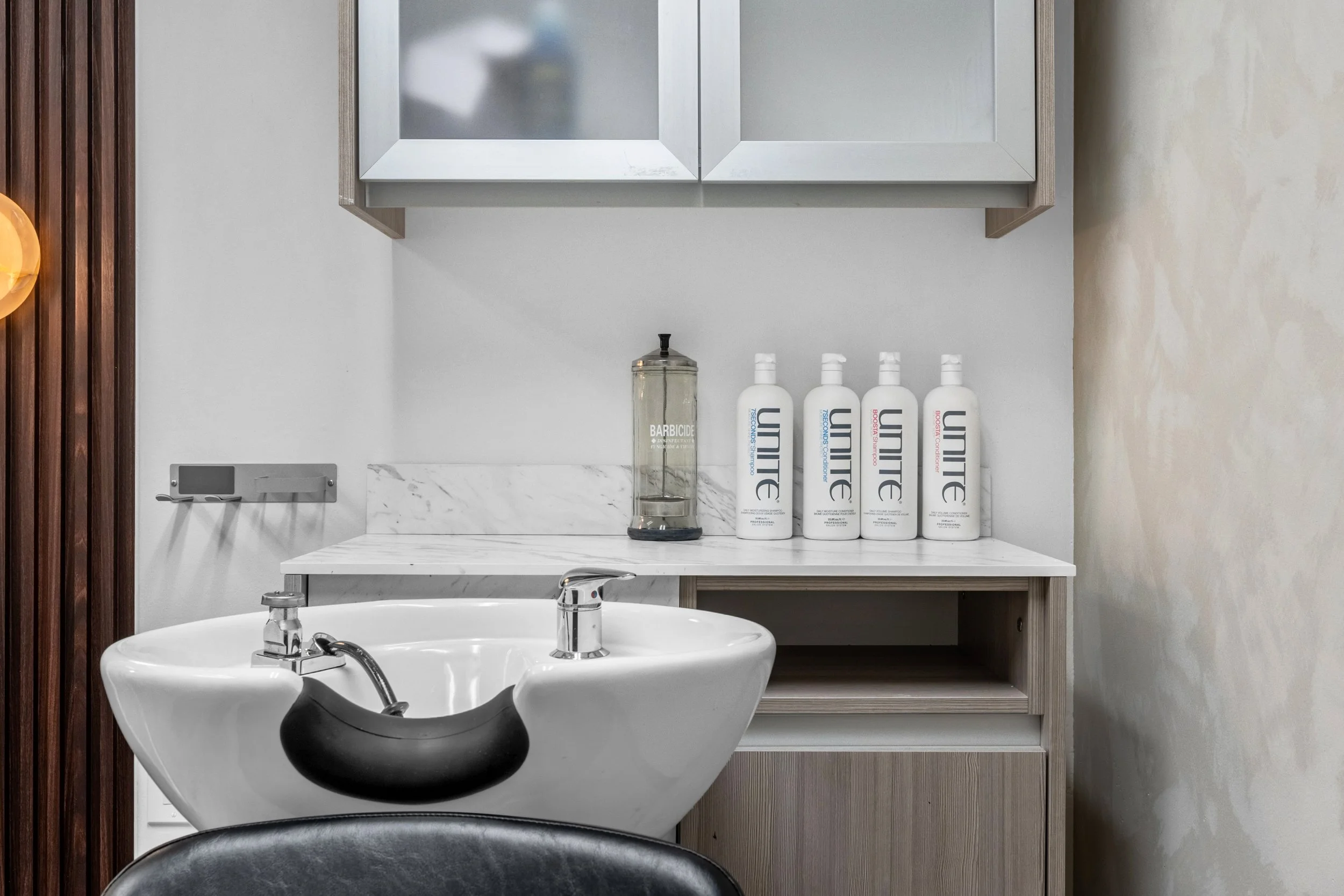Interior view of a salon or barber shop wash station with a white sink, black neck rest, and a countertop holding four bottles of Lume skincare products and a Barbicide jar.