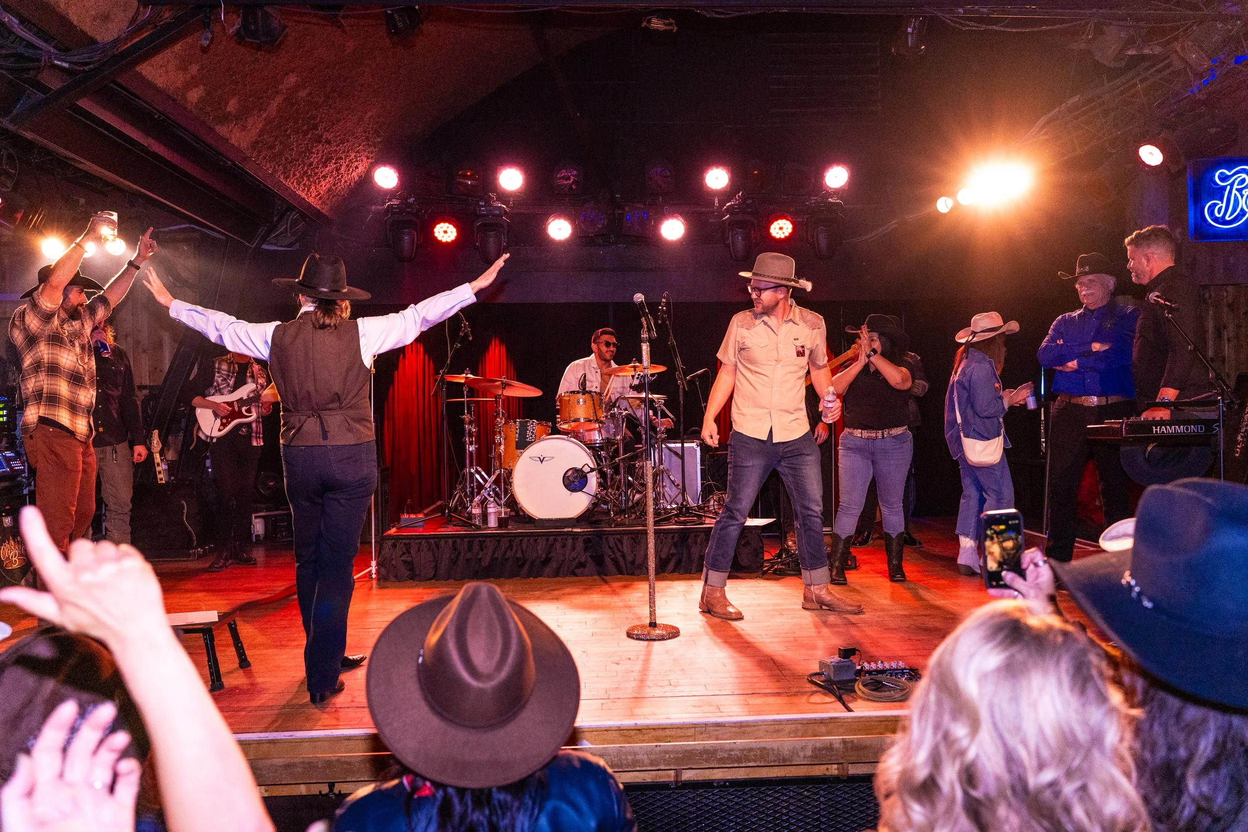 A lively stage performance with a band of musicians and dancers, including a drummer, guitarists, and singers, in a venue with colorful lighting and an engaged audience, some wearing cowboy hats.