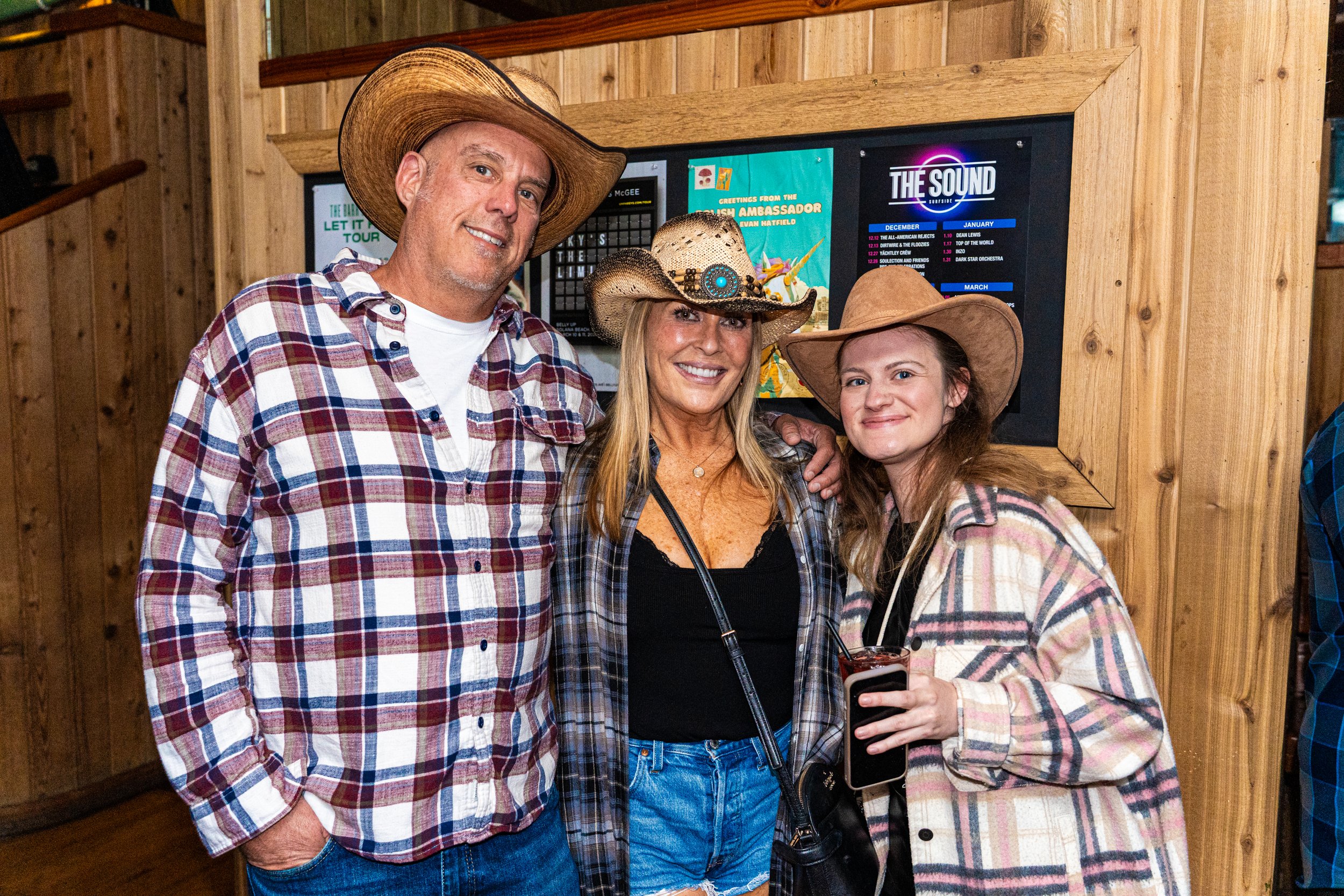 Three people wearing cowboy hats, standing together in front of a wooden wall with a television screen, smiling at the camera, in a rustic setting.