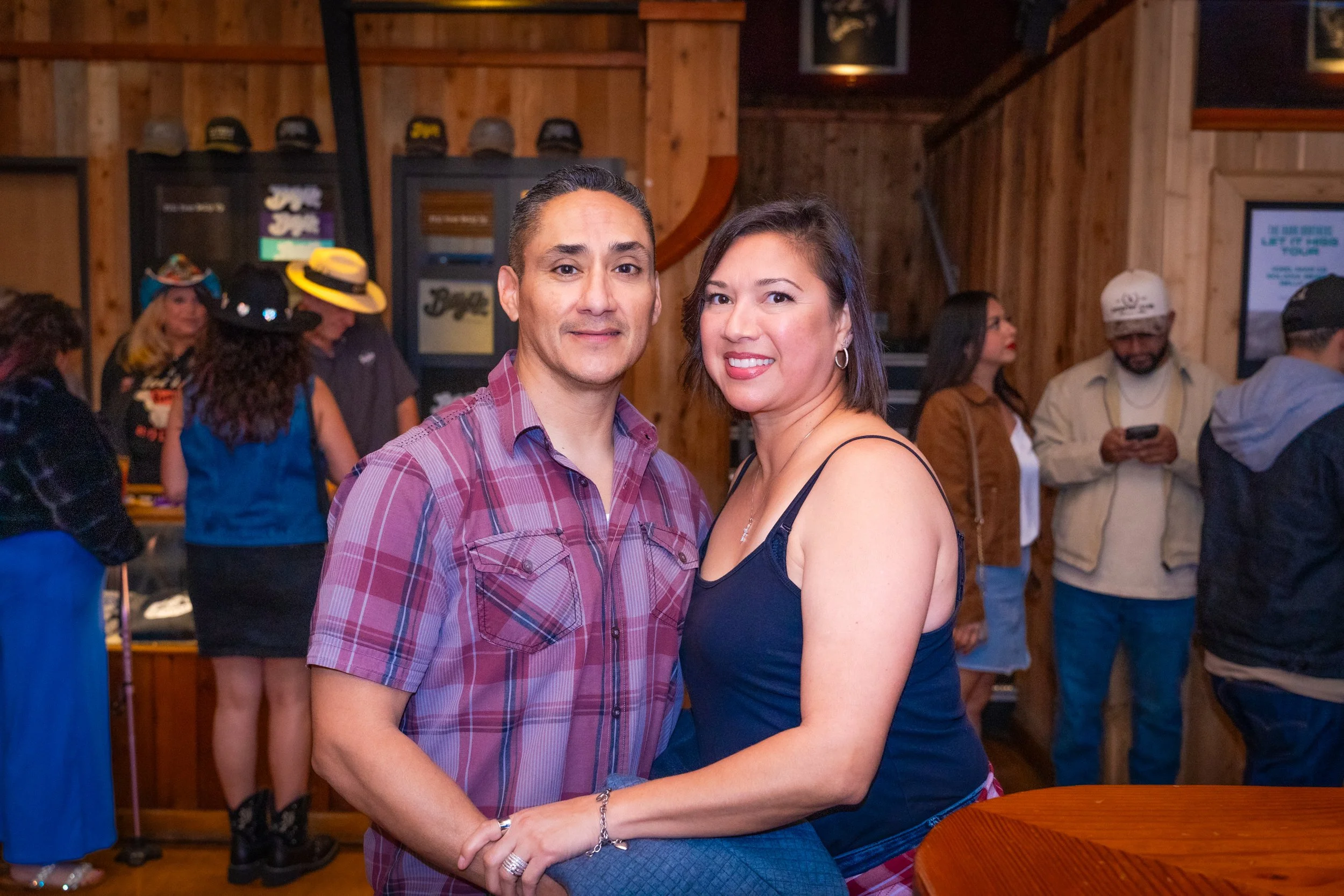 A man and woman posing together in a wooden-walled bar or restaurant. The man is wearing a red plaid shirt, and the woman is in a black spaghetti strap top. Several other people are in the background, some wearing cowboy hats, with wooden decor and f