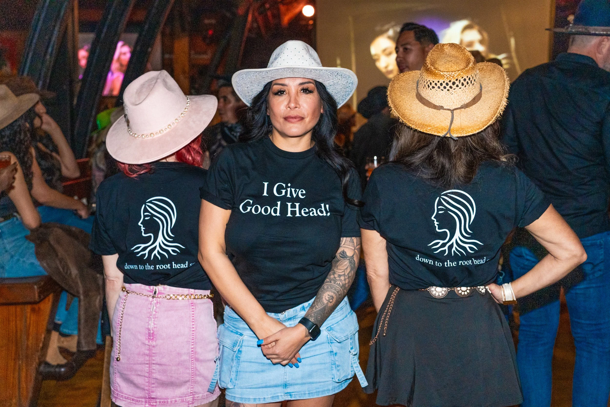 Three women at a social event wearing black T-shirts with slogans. The woman in the center wears a white hat and has long black hair with tattoos on her arm. The woman on the left wears a light pink hat and a pink skirt. The woman on the right wears 