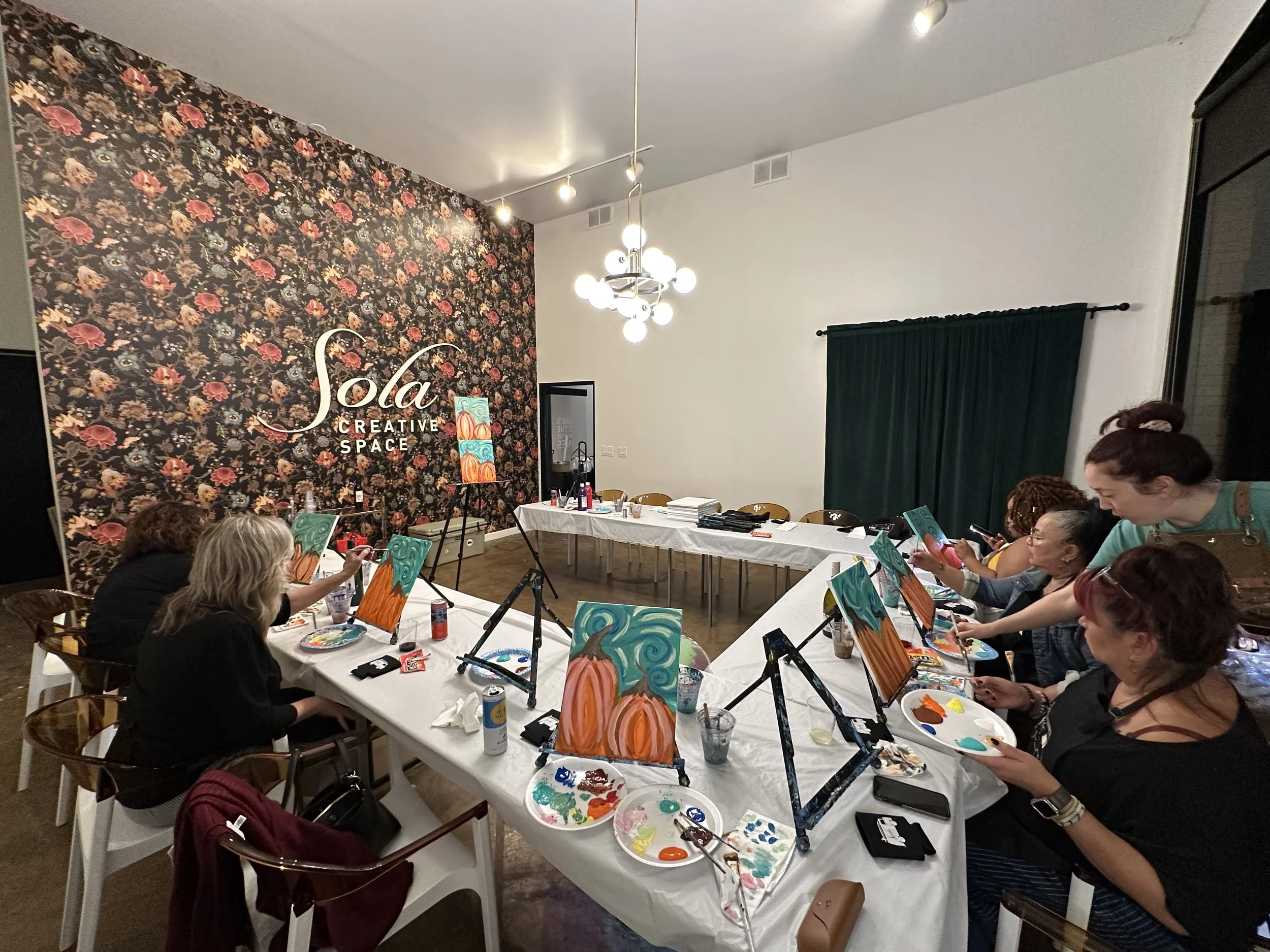 Group of women participating in a guided painting class at Solá Creative Space, with easels, paints, and canvases, in a decorated room with a floral accent wall.