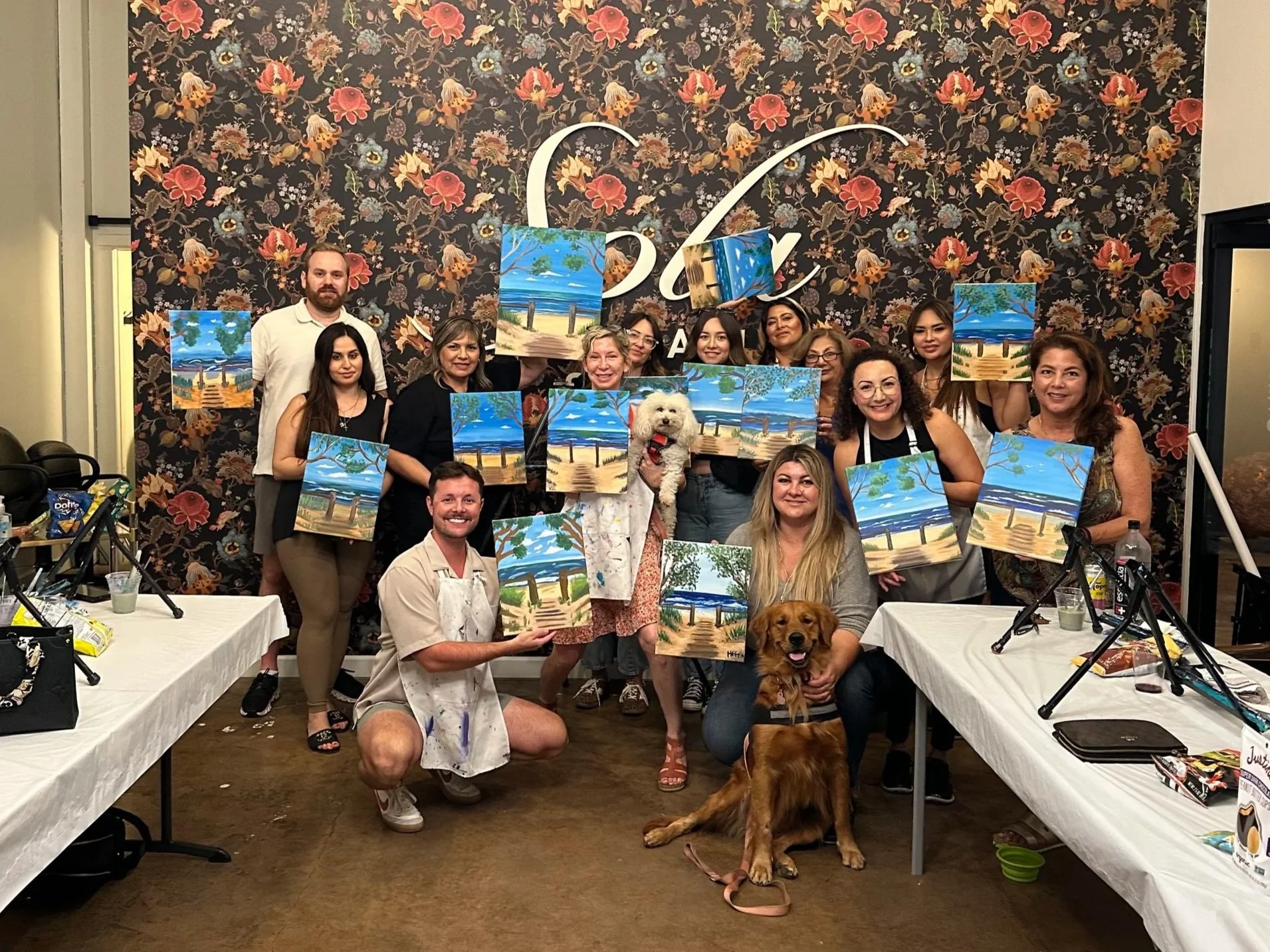 Group of people participating in a painting class or event, holding up finished landscape paintings featuring beachscapes, with dogs present, inside a room with floral wallpaper.