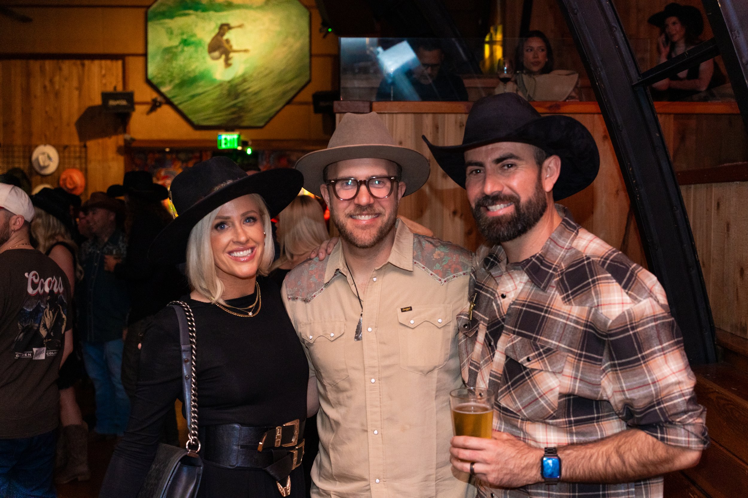 Three people smiling and dressed in cowboy-themed attire, including wide-brimmed hats, at a social event or party with a wooden interior and a crowd in the background. The person on the right is holding a glass of beer.