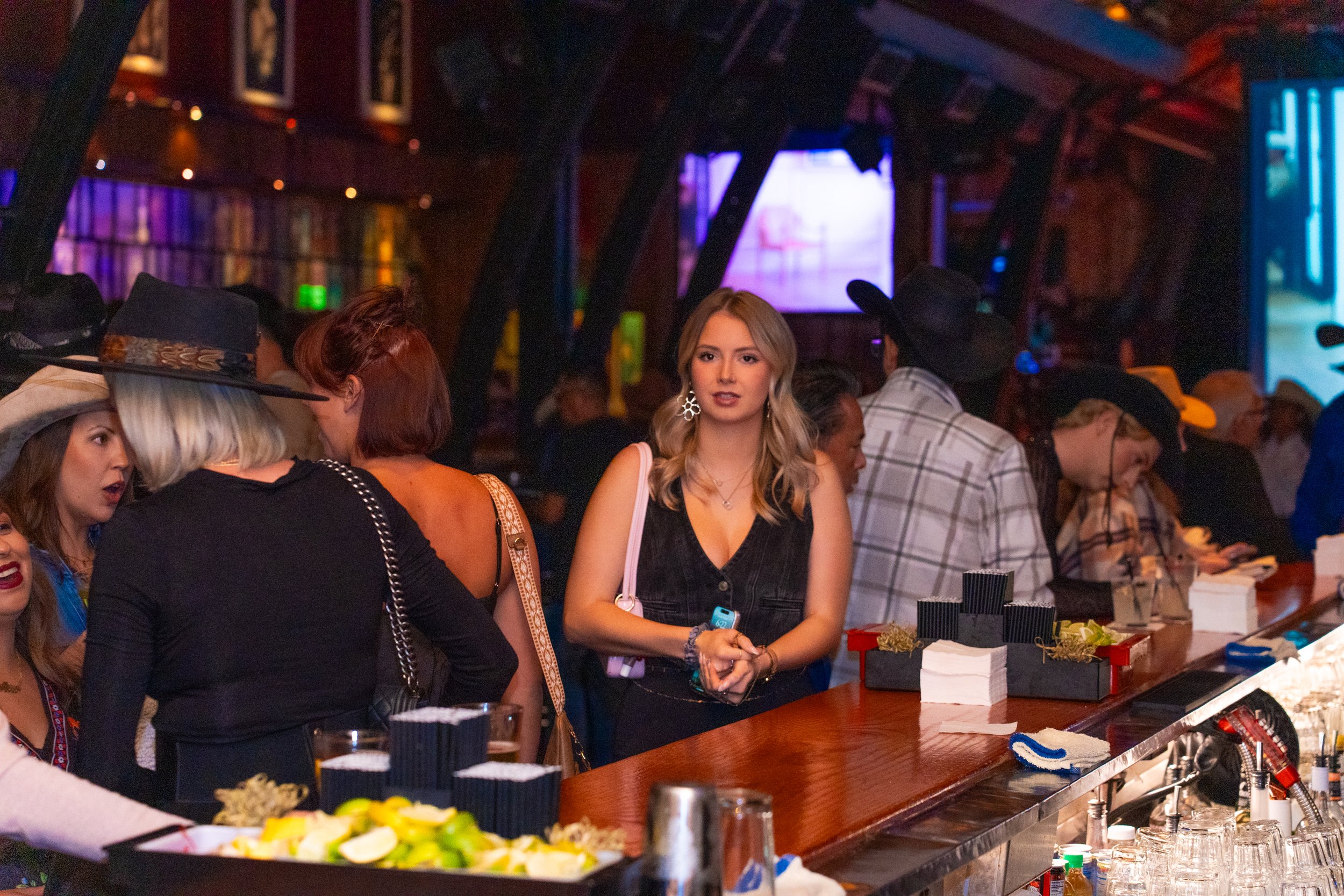 People at a bar, some wearing cowboy hats, in a dimly lit venue with colorful lighting and a large screen in the background.
