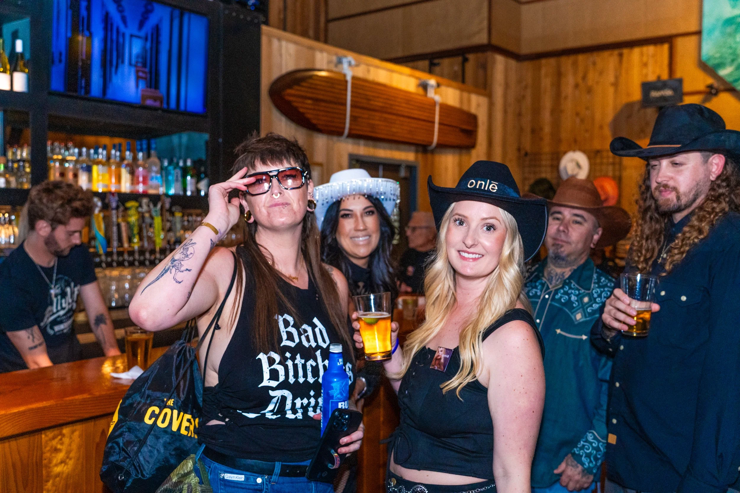 Group of people wearing cowboy and Western-themed costumes, enjoying drinks at a bar.