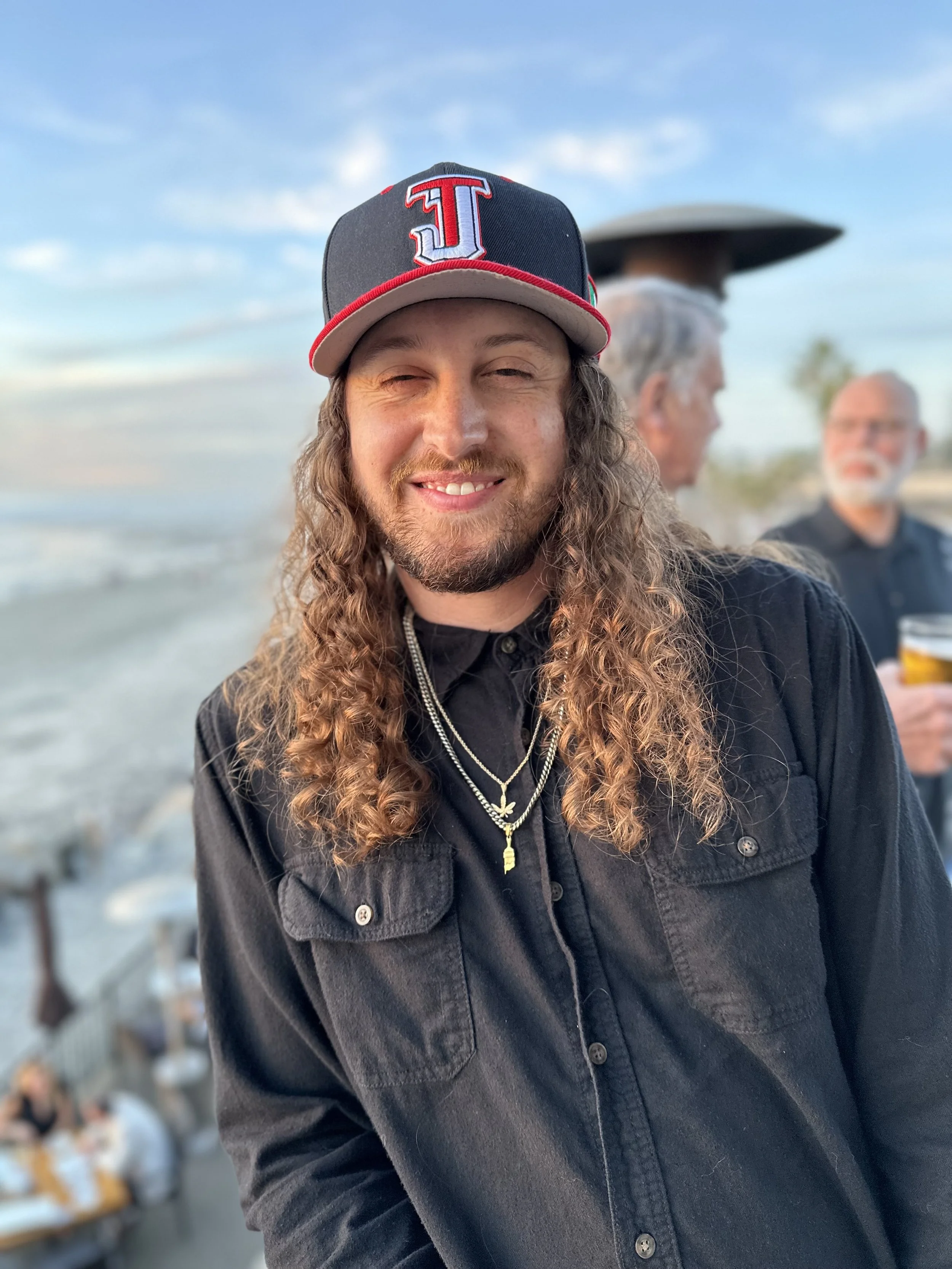A man with long curly hair and a beard smiling at the camera, wearing a black shirt and a baseball cap with a red and gray 'J' logo, at a beach with other people in the background.