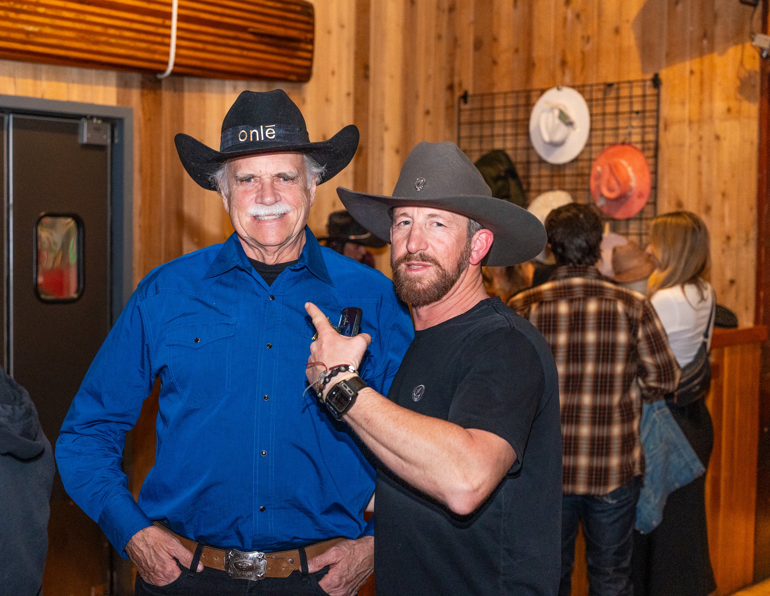 Two men wearing cowboy hats pose together indoors, with a wooden wall and hats displayed on a grid in the background.
