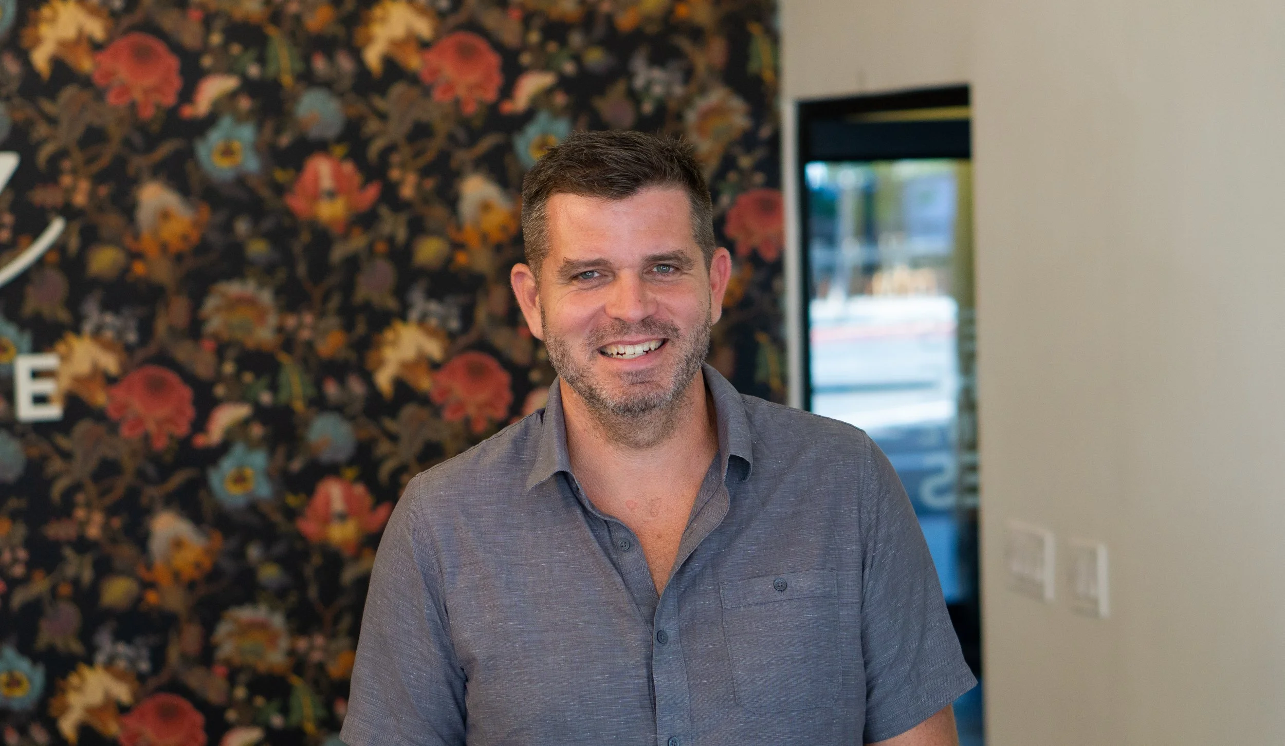 A smiling man with short brown hair and a beard, wearing a gray button-up shirt, standing indoors with a floral patterned wall in the background.