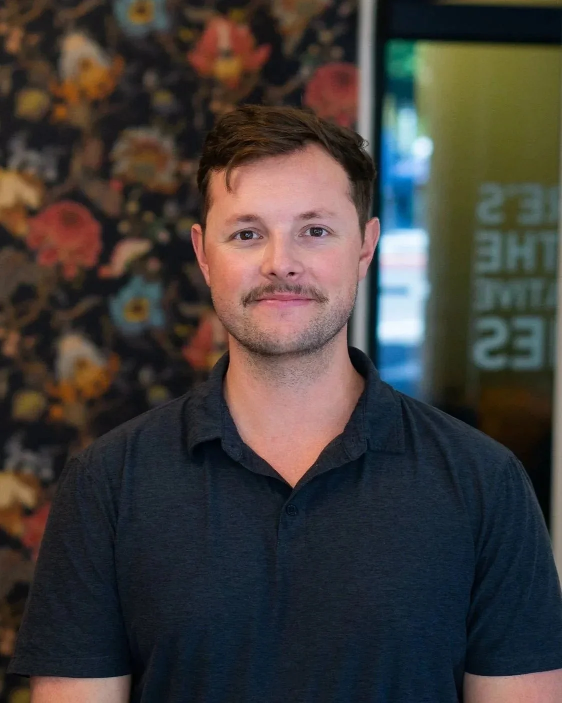 A young man with short brown hair and a trimmed beard, wearing a black collared shirt, standing indoors with a floral patterned wall behind him and a glass window to his right.