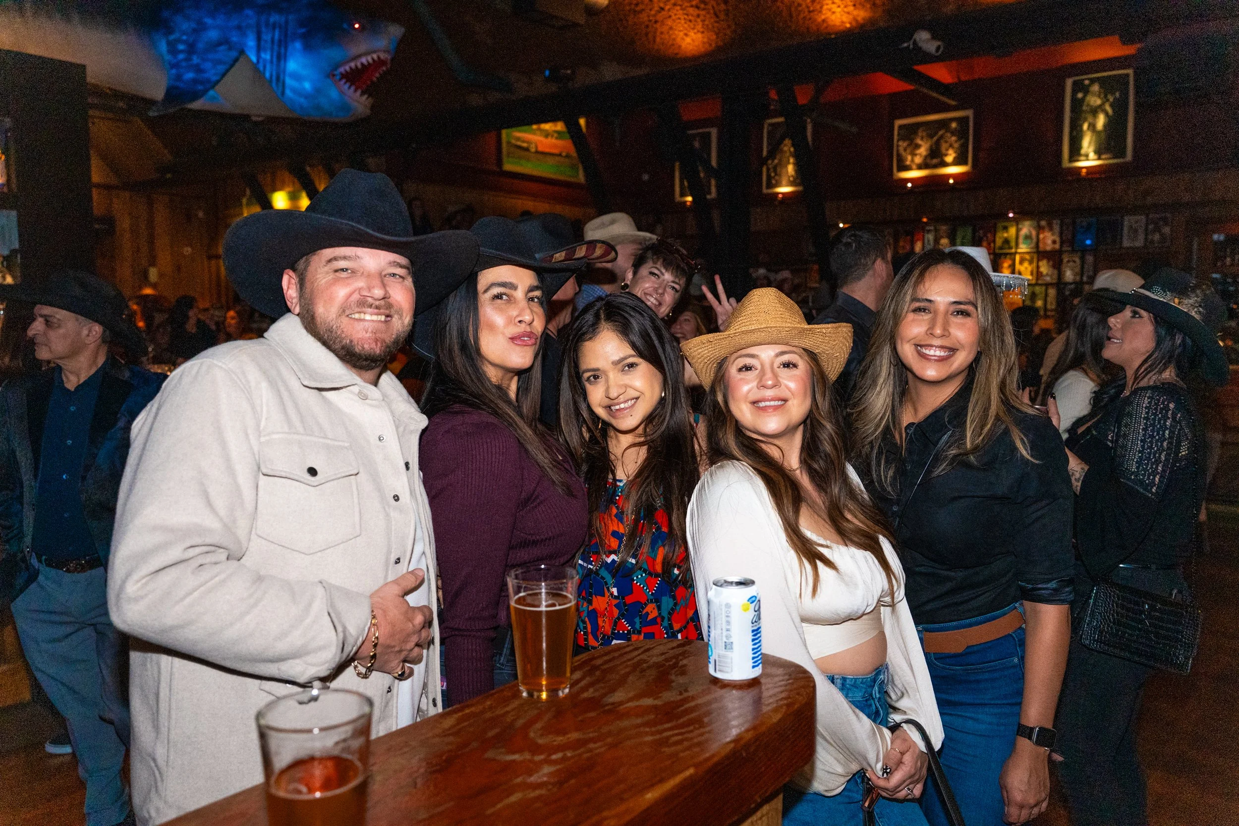Group of people at a bar wearing cowboy hats, smiling, with drinks on the counter, with a shark decoration on ceiling and framed pictures on wall in background.