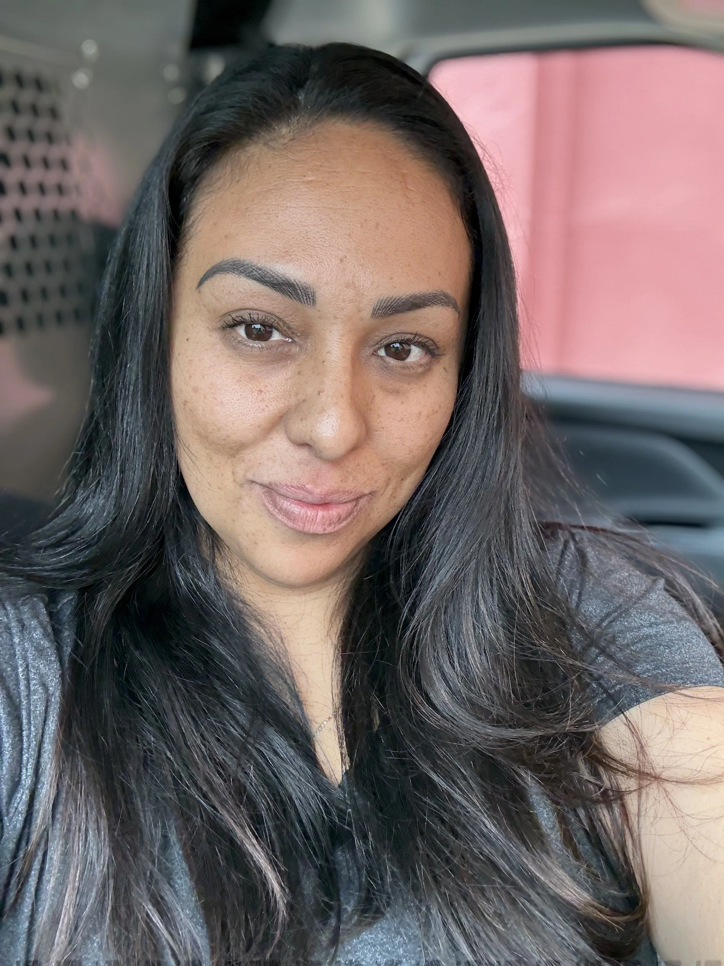 A woman taking a selfie inside a vehicle, smiling with long dark hair, brown eyes, and wearing a gray top.
