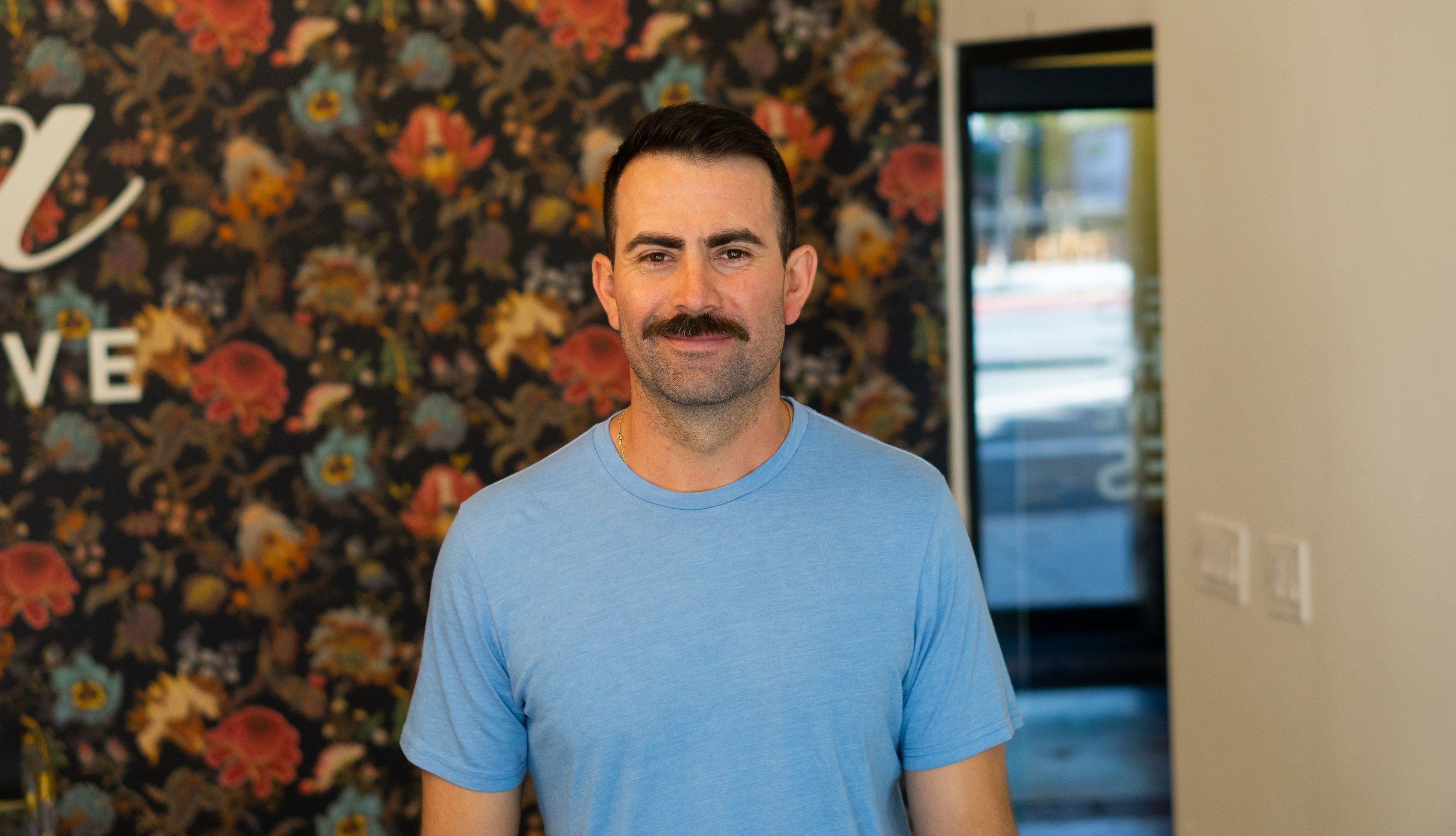 A man with dark hair and a moustache wearing a light blue t-shirt, standing indoors in front of a floral wall with the word 'LOVE' partially visible behind him.