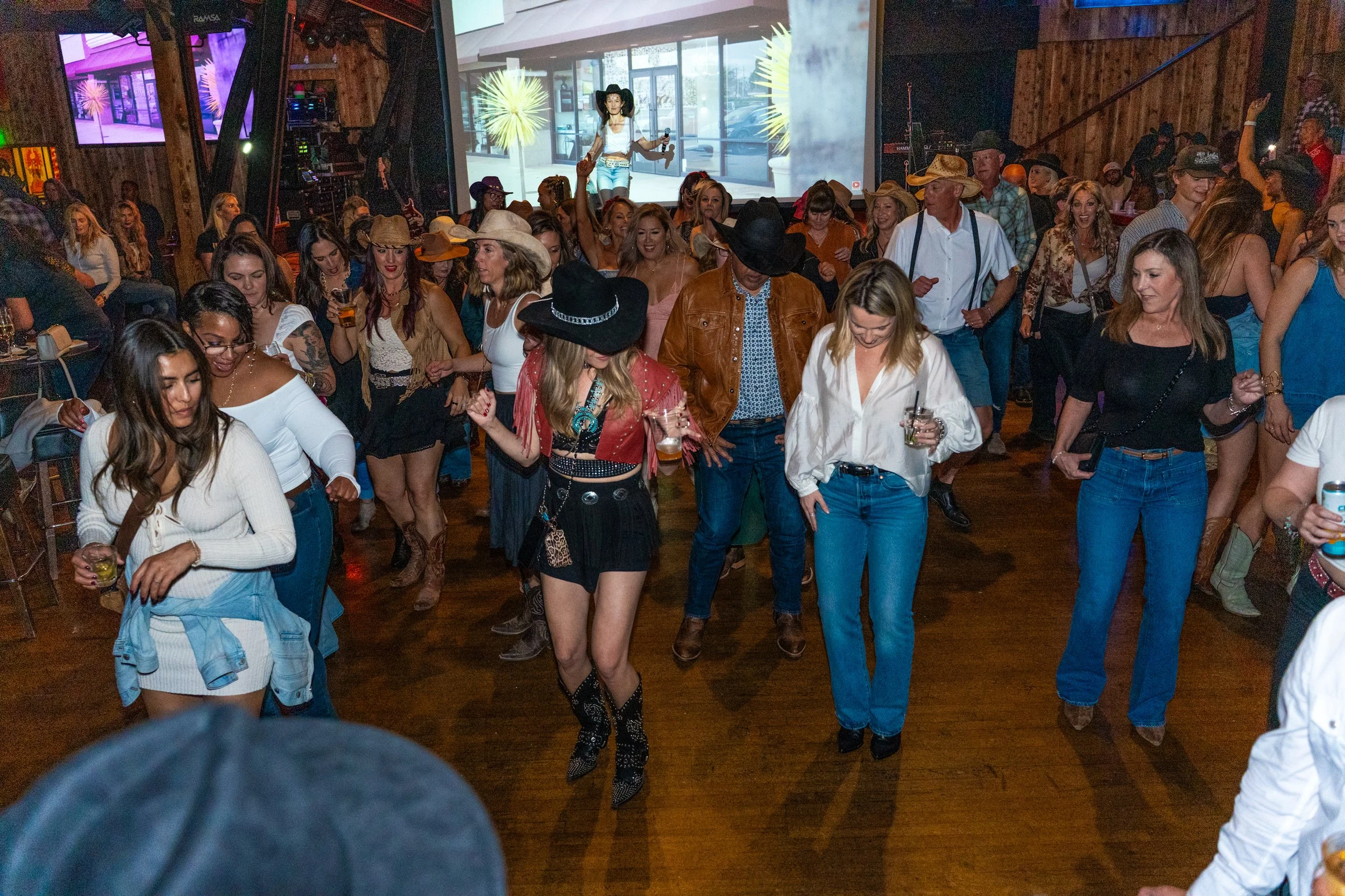 People line dancing in a rustic bar or club with wood floors and walls, wearing country or western attire, some with cowboy hats, and enjoying drinks.