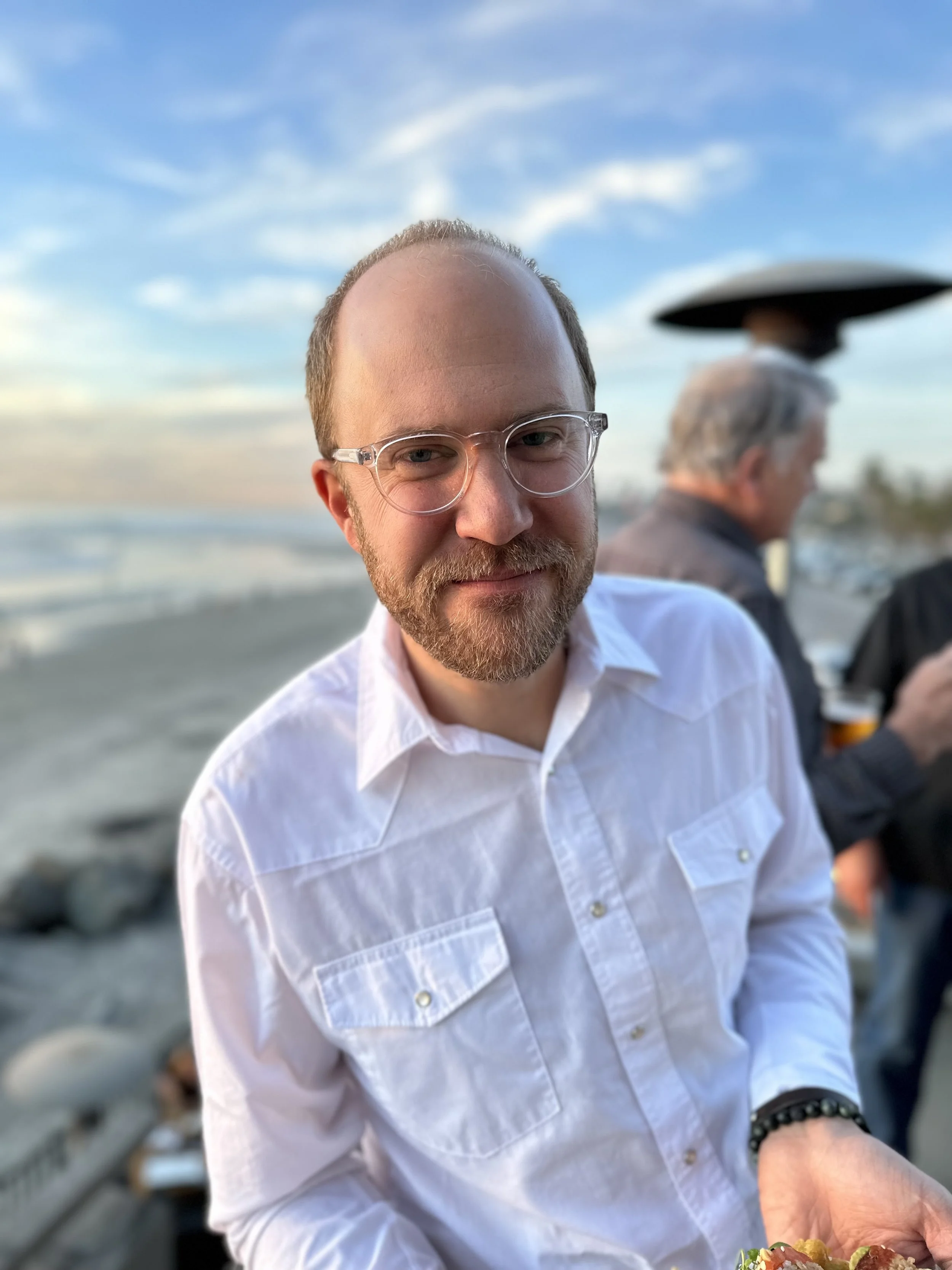 A man wearing a white shirt and glasses, smiling at camera, at the beach during sunset with people in the background, one holding an umbrella.