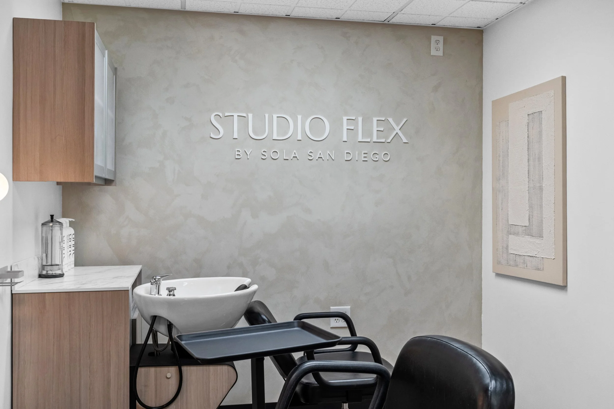 Interior of a hair salon with a sign on the wall reading 'STUDIO FLEX BY SOLA SAN DIEGO', a white ceramic sink with a black hairdryer on it, a wooden cabinet with a sanitizer dispenser, two black chairs, and an abstract painting on the wall.