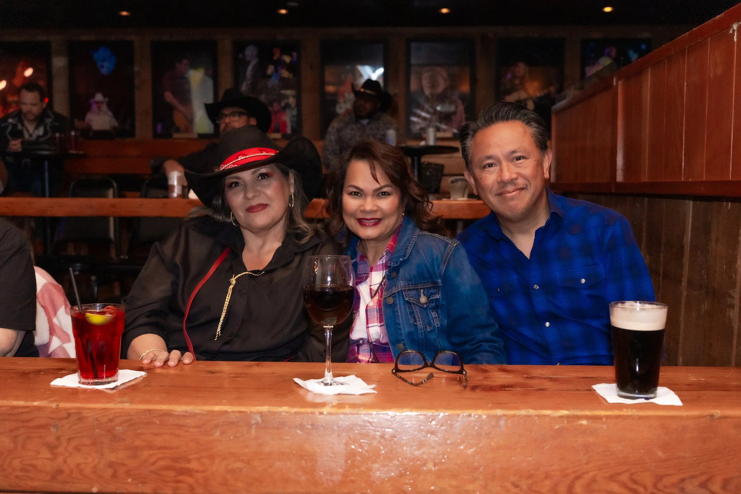 Three people sitting at a bar or restaurant table, smiling and enjoying drinks, with a background of framed pictures and other patrons.