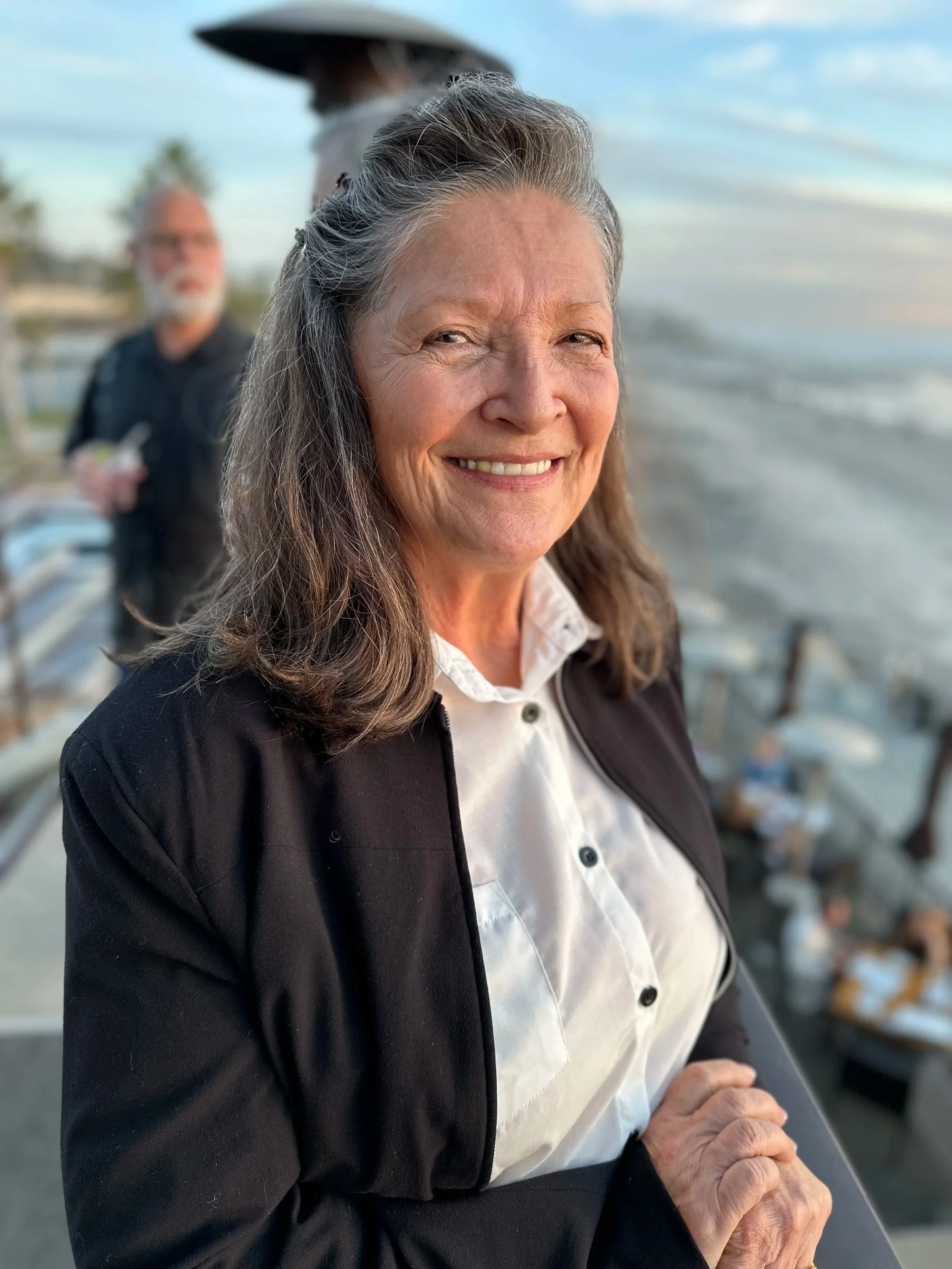 A smiling elderly woman with gray hair in a black jacket and white shirt standing on a deck near the water during sunset, with two out-of-focus men in the background.