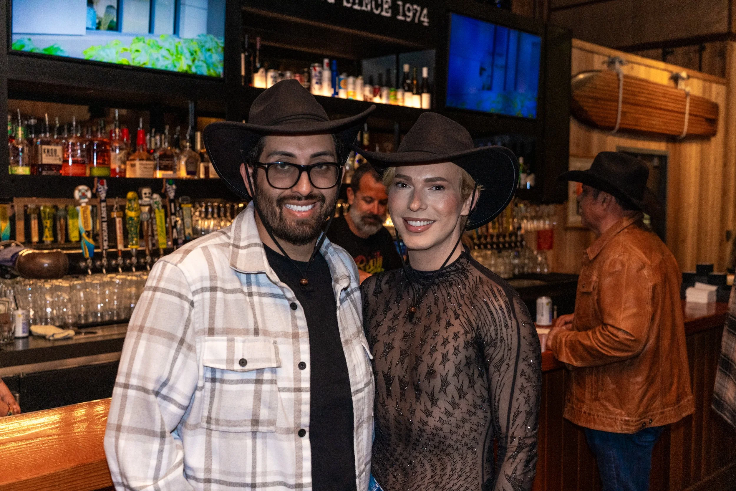 A man and woman dressed as cowboys smiling at the camera at a bar. The man is wearing glasses, a plaid shirt, and a cowboy hat. The woman is wearing a sheer, star-patterned top and a cowboy hat. There are bottles and glasses behind them at the bar, w