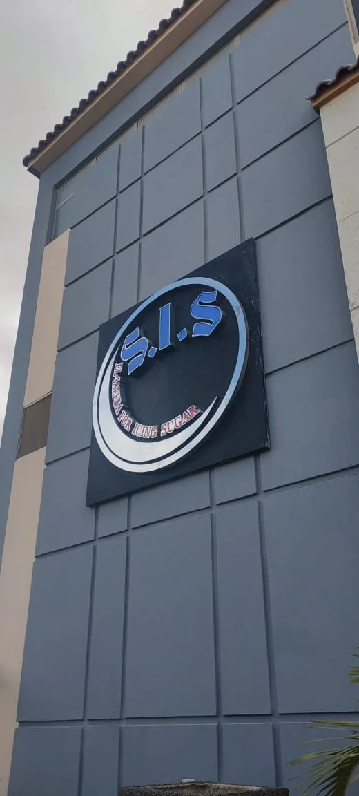 A building exterior with a sign that reads "S.I.S" in large blue letters and a tagline underneath that says "Sweetening Your Icing with Sugar" in red text, all mounted on a gray wall.