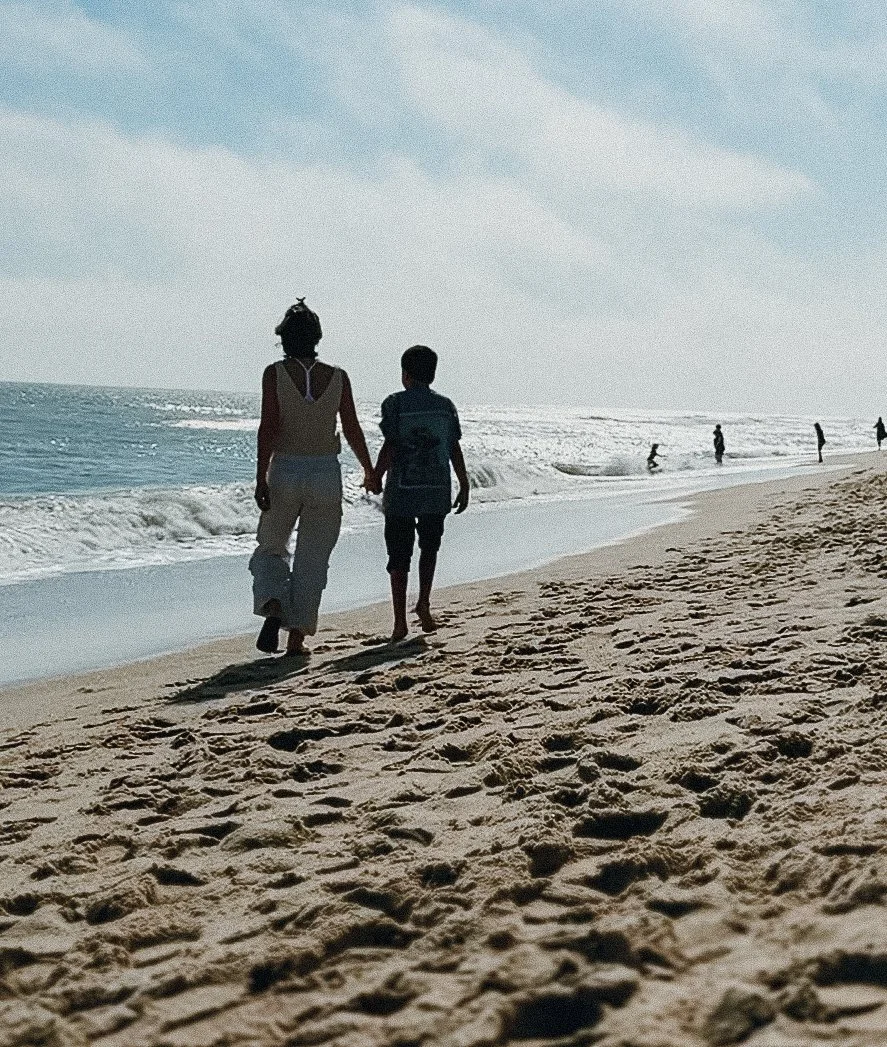 A woman and a child holding hands walking along the beach by the ocean during daytime.