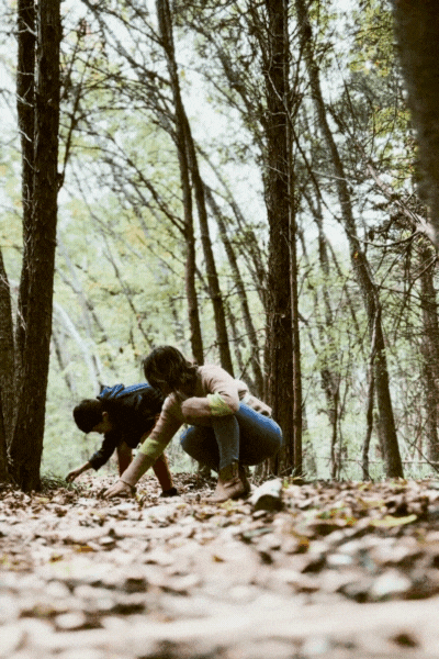 Two children playing in a forest, crouched on the ground among fallen leaves.