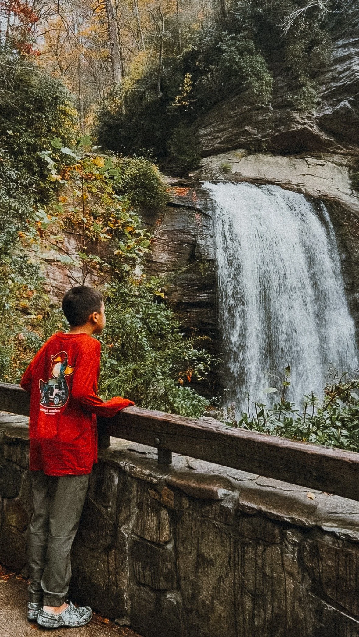 A boy in a red jacket stands at a stone and wood railing, gazing at a waterfall in a forest with fall foliage.