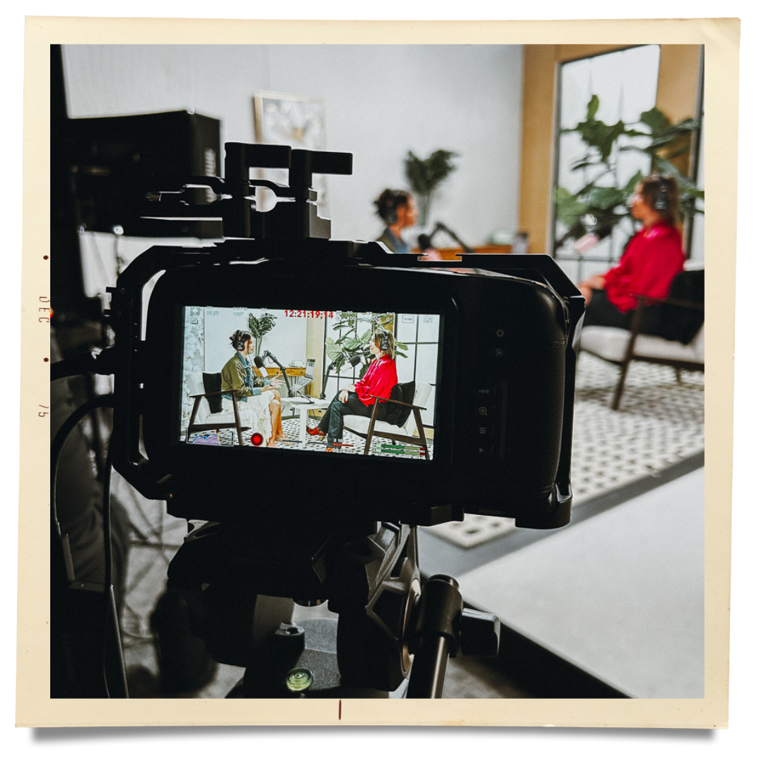A camera recording a podcast interview with two women sitting on chairs in a modern room, with one woman in a red shirt and the other in a green jacket, and a large window with plants in the background.