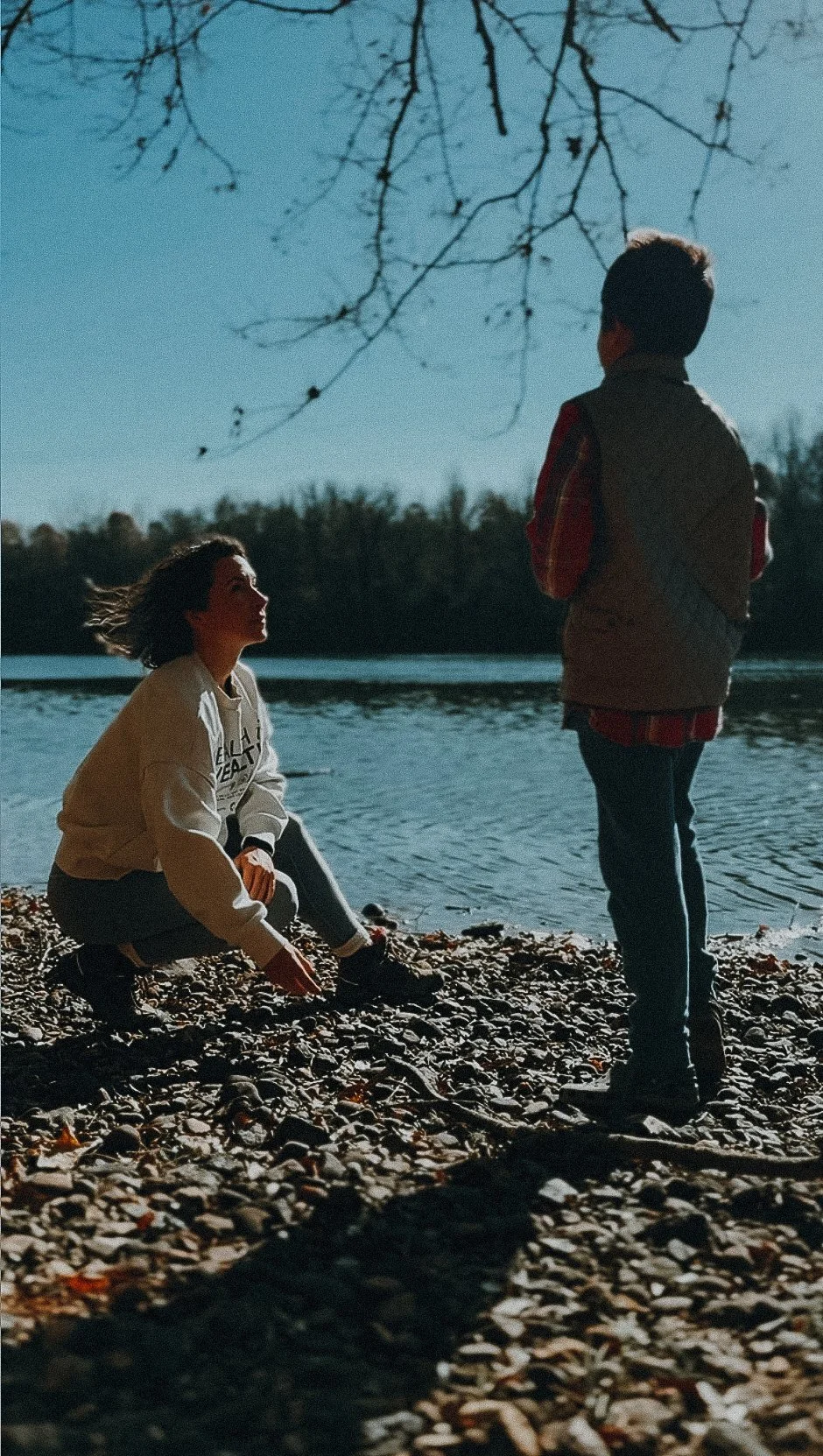 A woman sitting on rocky beach by water, facing a boy standing nearby, with trees in the background, during dusk or dawn.