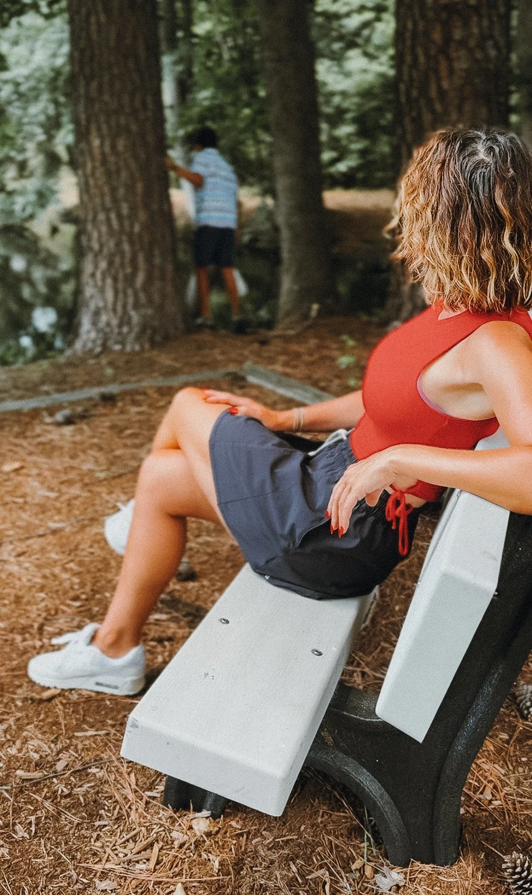 A woman sitting on a park bench looking towards a young boy climbing a tree in a forested area.