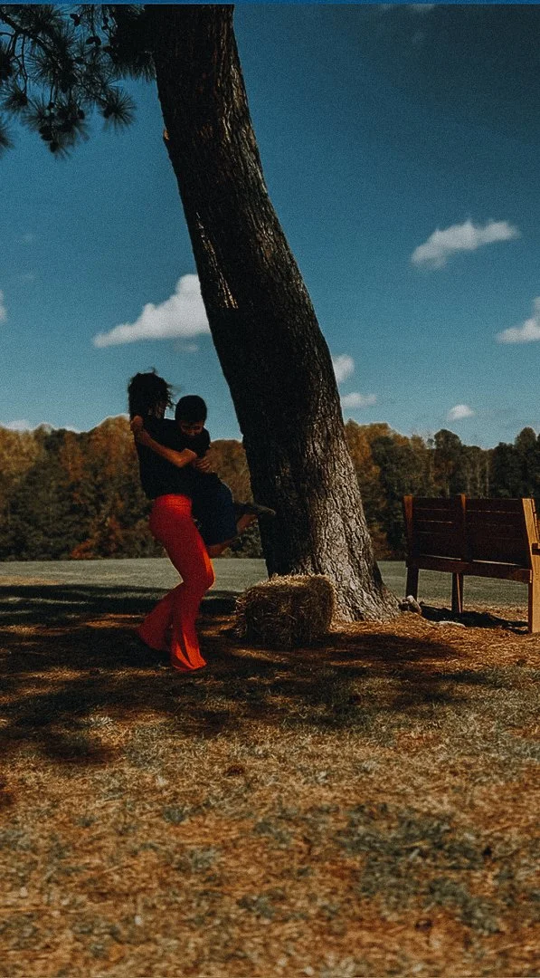 A person in red pants holding a child in black near a large tree in a park with a bench and a forest in the background on a sunny day.