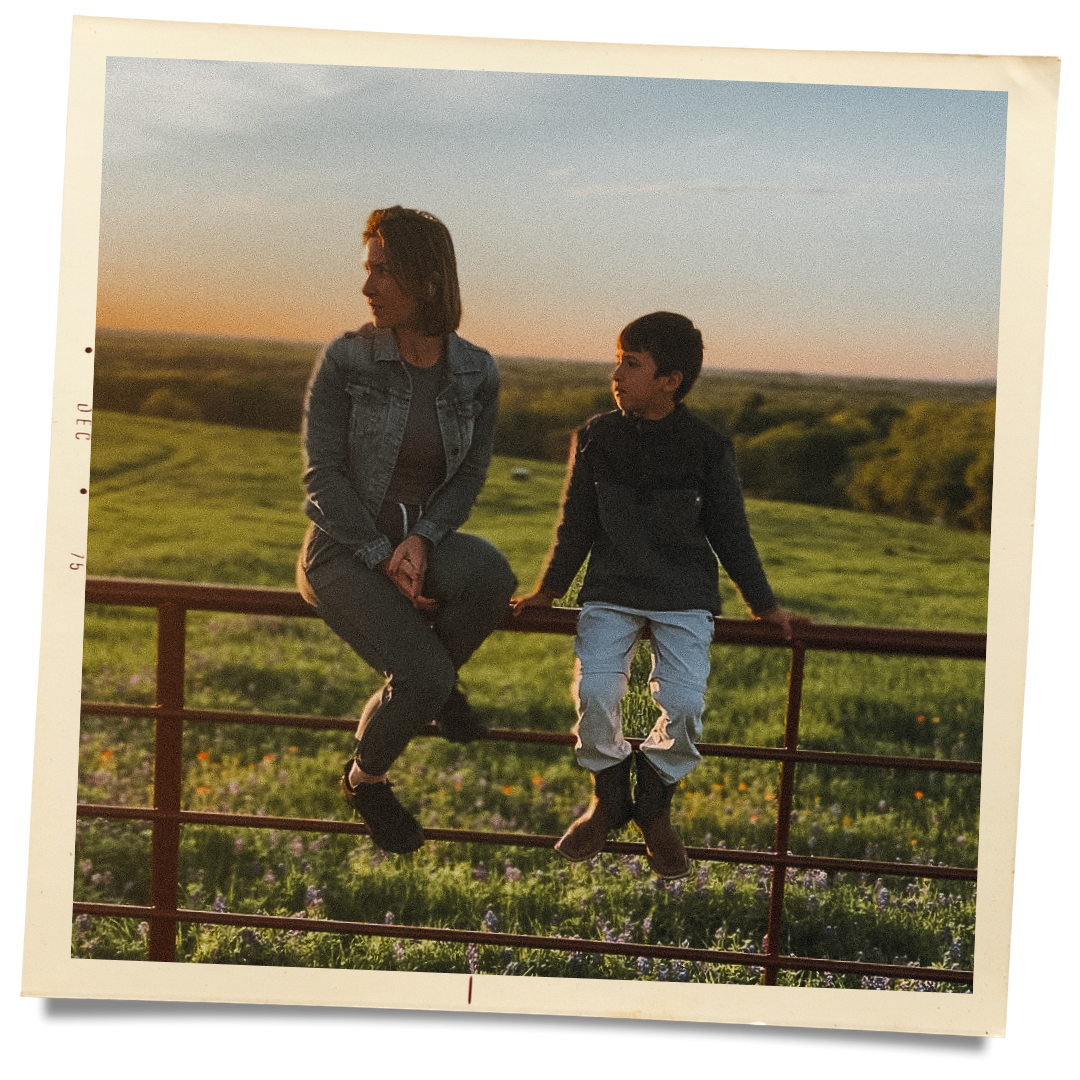 A woman and a young boy sitting on a metal fence in a rural field during sunset, with green grass and trees in the background.
