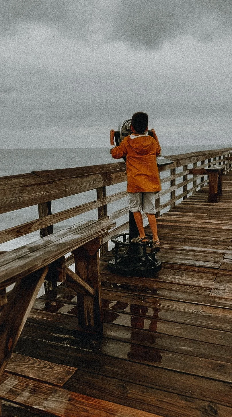 A boy in an orange jacket and shorts stands on a wet wooden pier, looking through a coin-operated binocular viewer at the overcast sky and ocean.