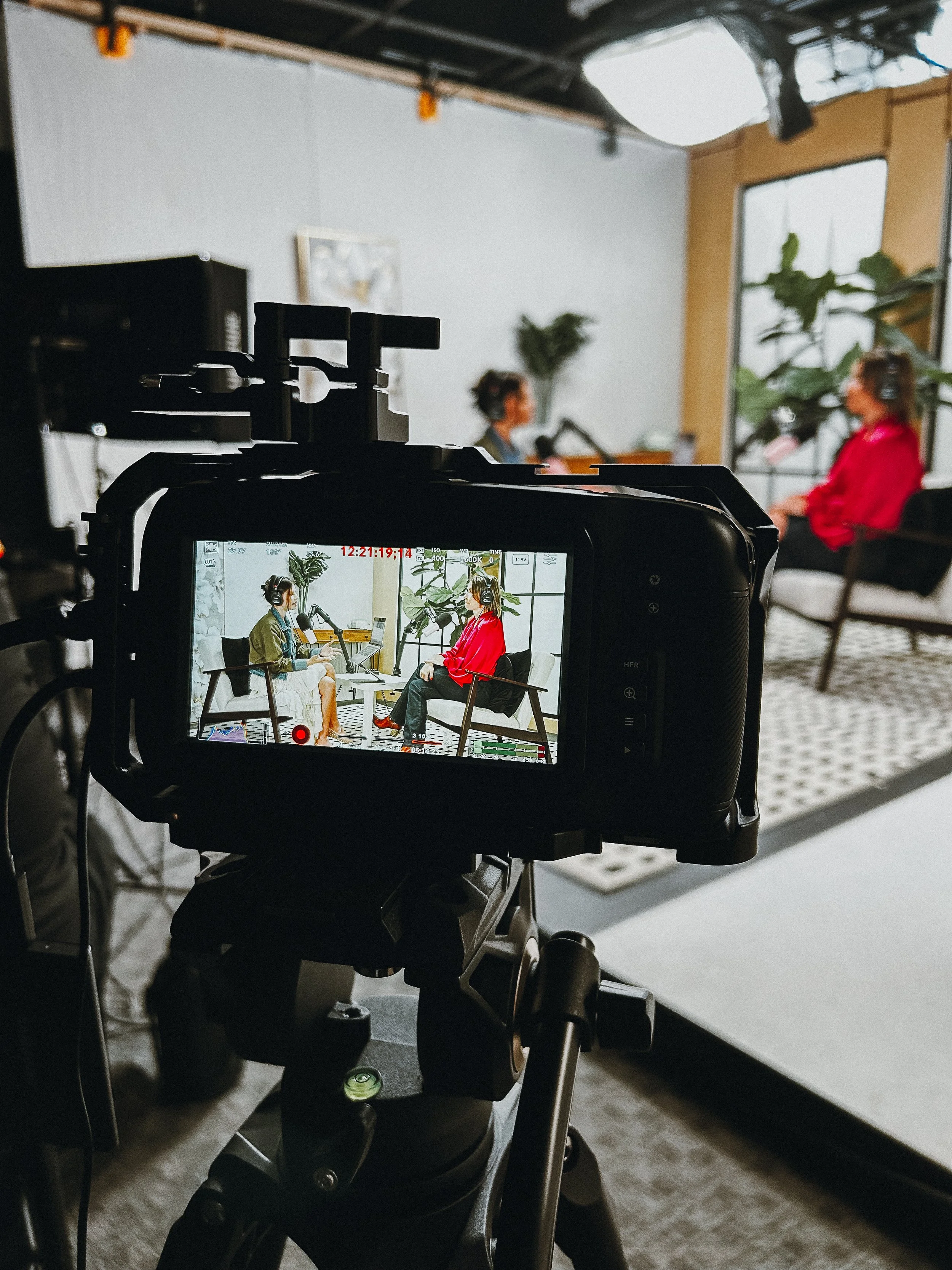 Two women recording a podcast in a studio, with one woman in a red jacket and the other in an olive green jacket, visible on a camera screen.