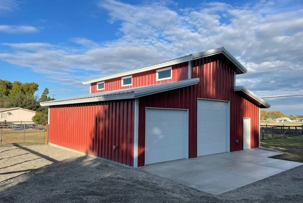Red metal barn with white trim, two garage doors, a side door, and a small window on the upper level, set on a concrete and gravel surface with a rural background.